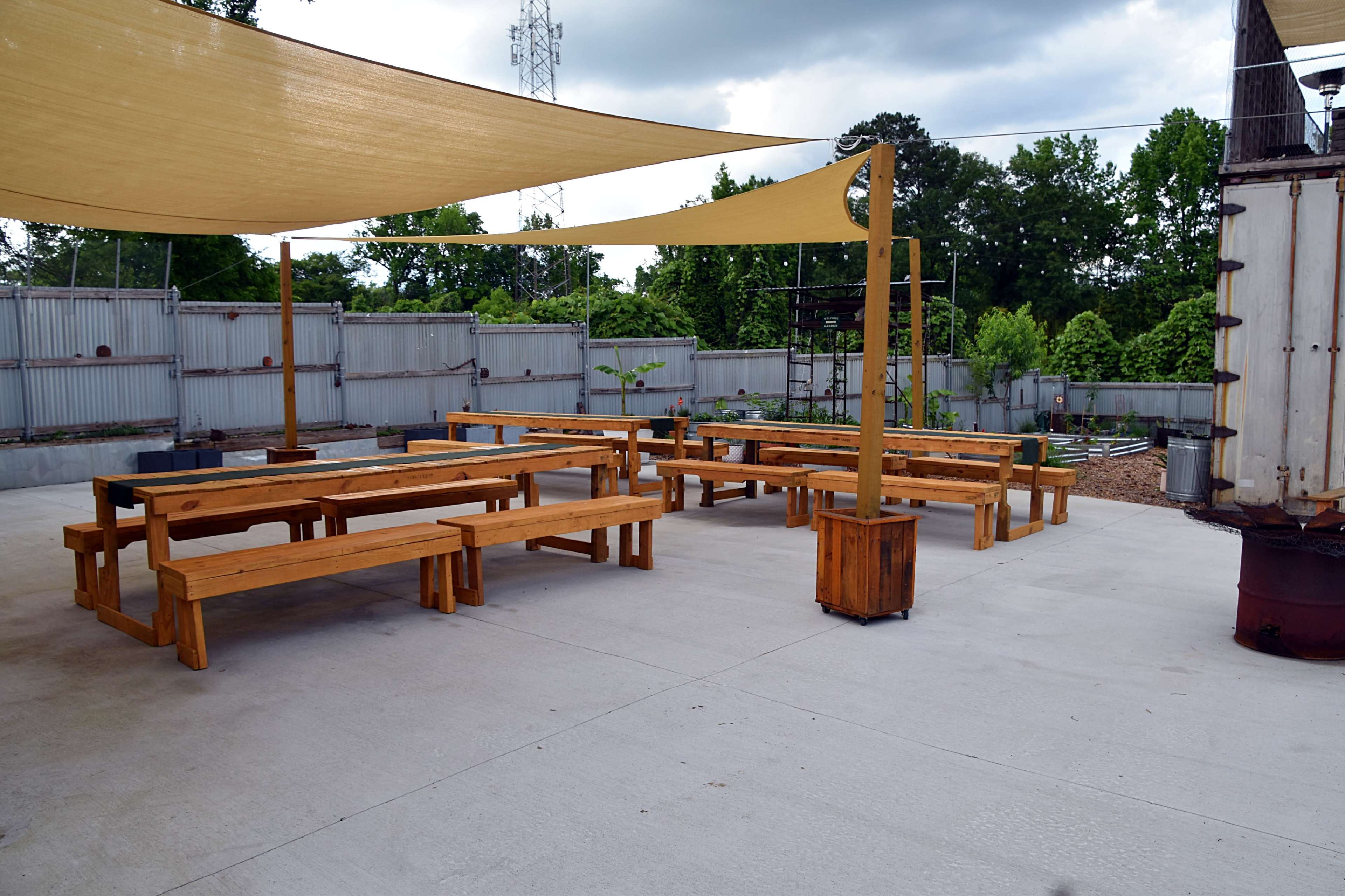 The image shows an outdoor seating area with wooden benches and tables shaded by large fabric canopies, surrounded by greenery and a gray fence.