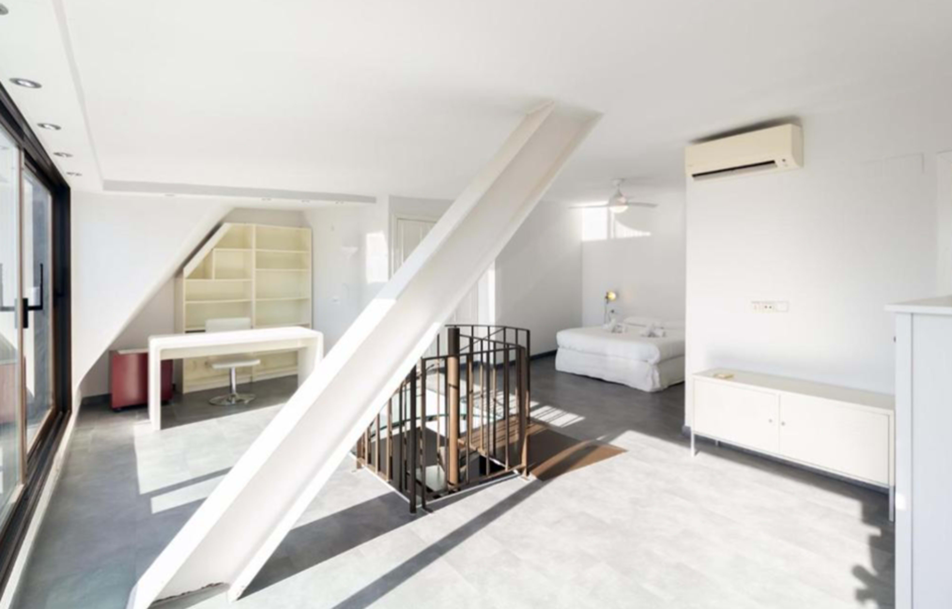 The image shows a modern, open-concept living space with a white color scheme, featuring a staircase, a workspace with a desk, and a seating area.