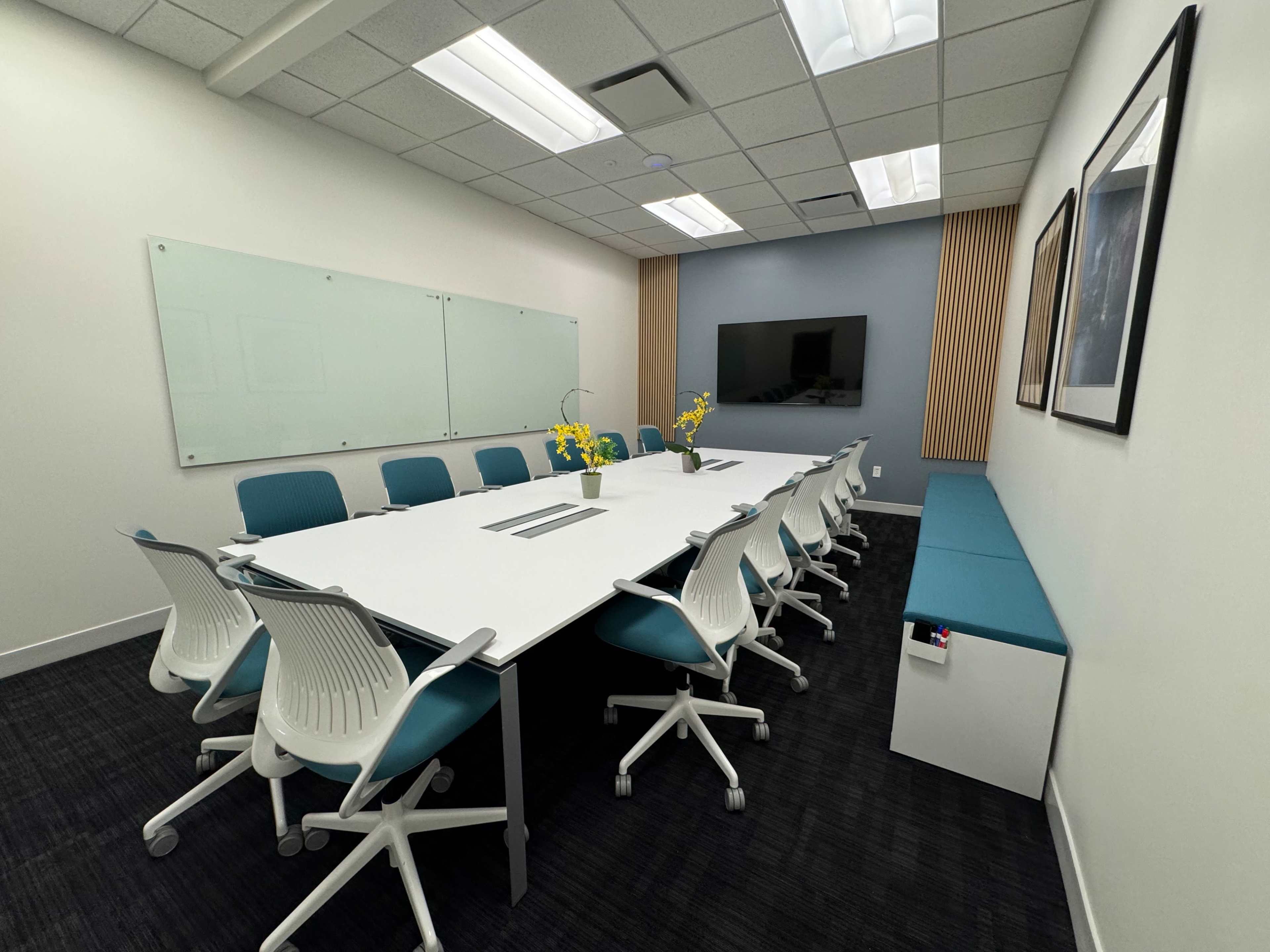 A modern conference room featuring a large white table surrounded by blue and white chairs, with a wall-mounted television and a whiteboard.