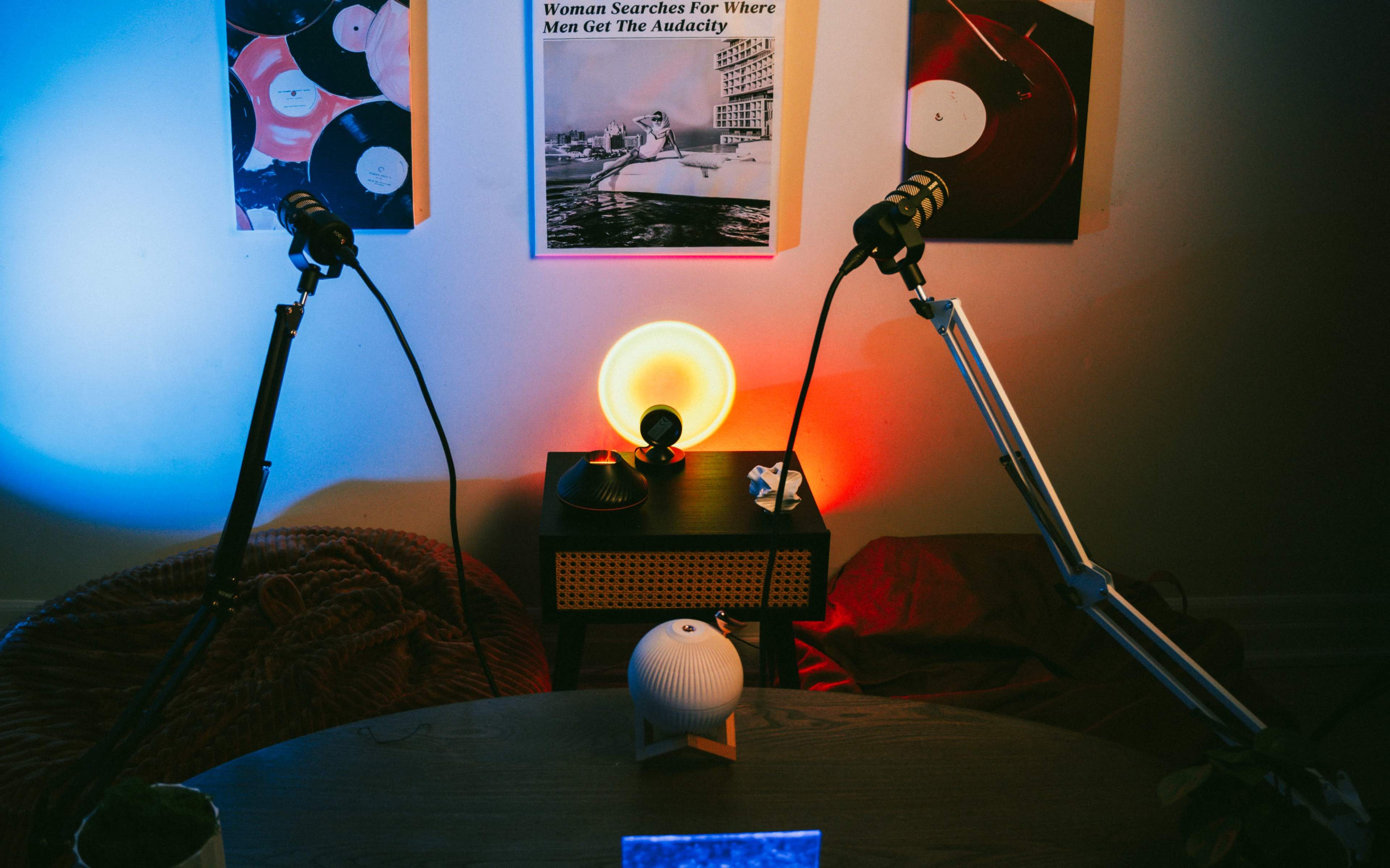 The image features a recording setup with two microphones on adjustable stands, a round wooden table, and ambient lighting in blue and red, alongside a stylized lamp and various decorative items.