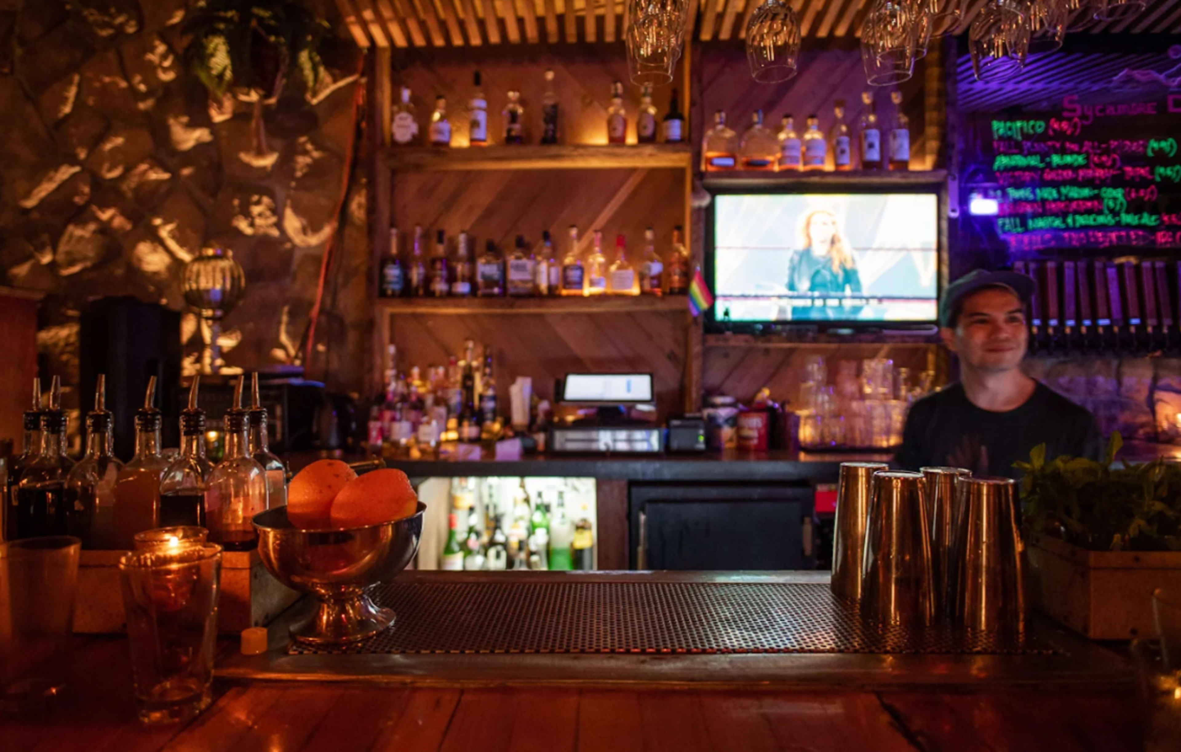 A bar scene features various bottles lined on shelves behind the counter, with a bartender preparing drinks and a television displaying a program in the background.
