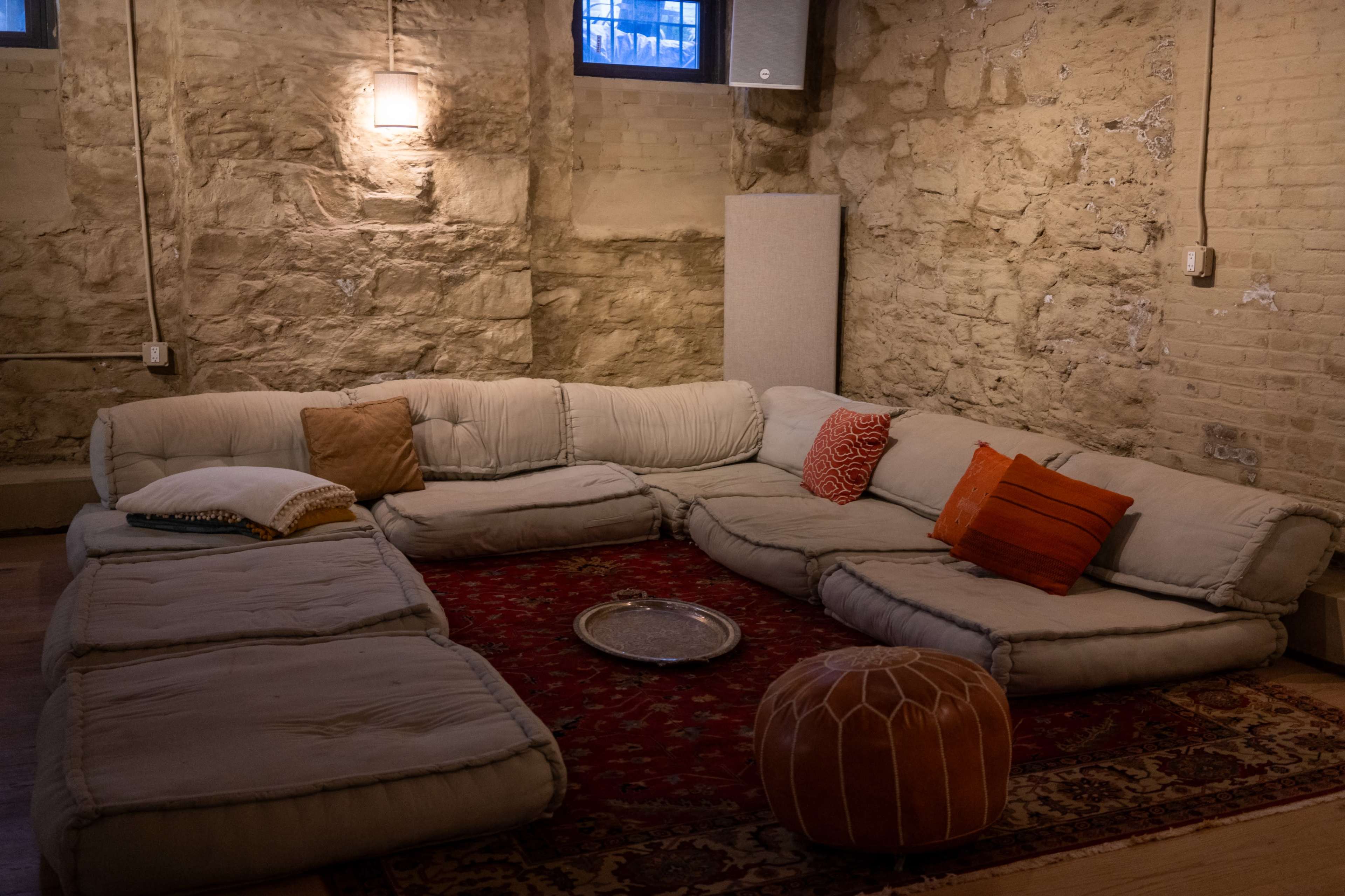 The image shows a cozy seating area with a large sectional couch, several cushions, and a round pouf on a red patterned rug against a stone wall.