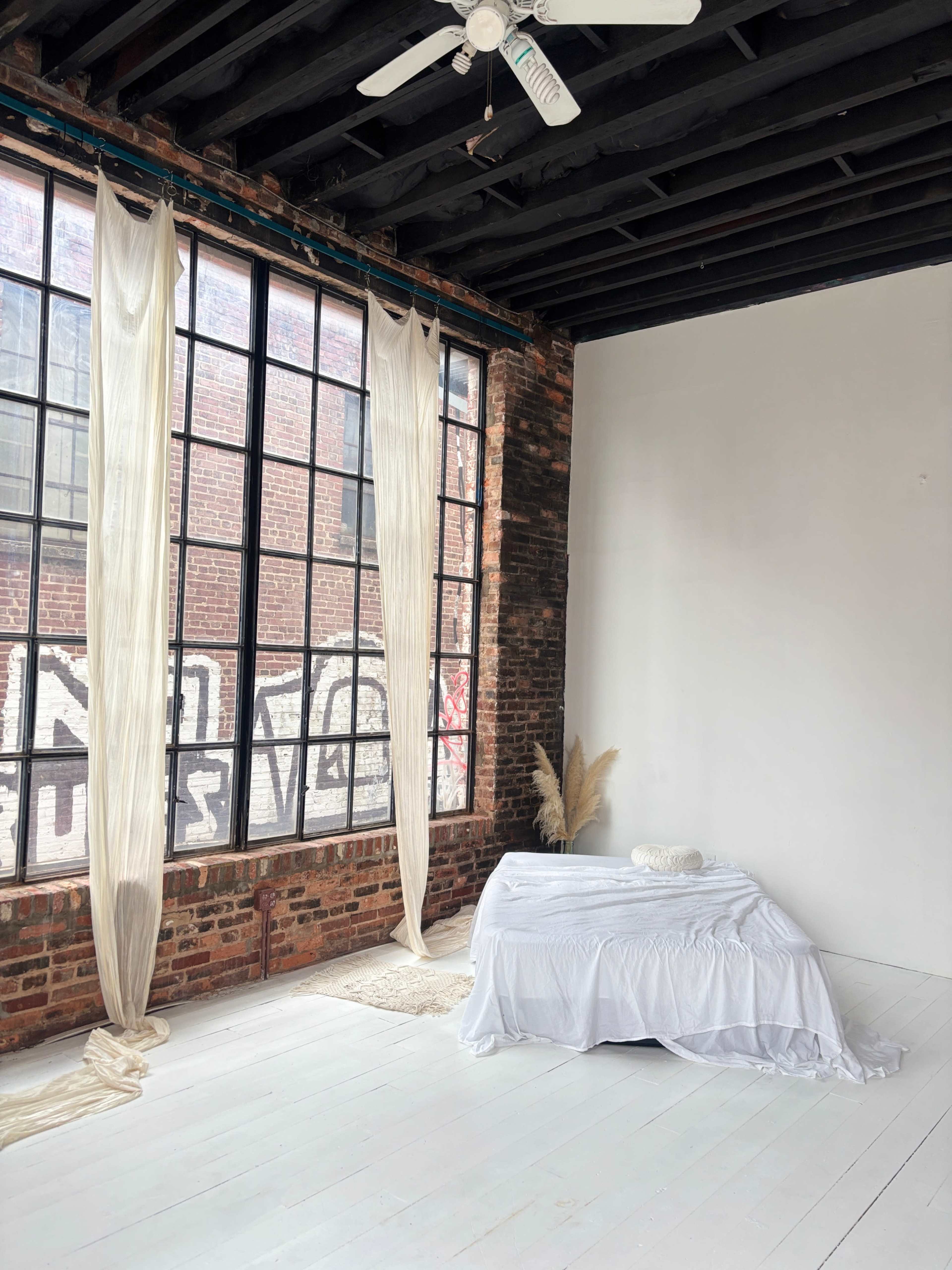 The image shows a minimalist bedroom with a white bedspread, large windows with sheer curtains, brick walls, and a simple decorative touch of dried plants in the corner.