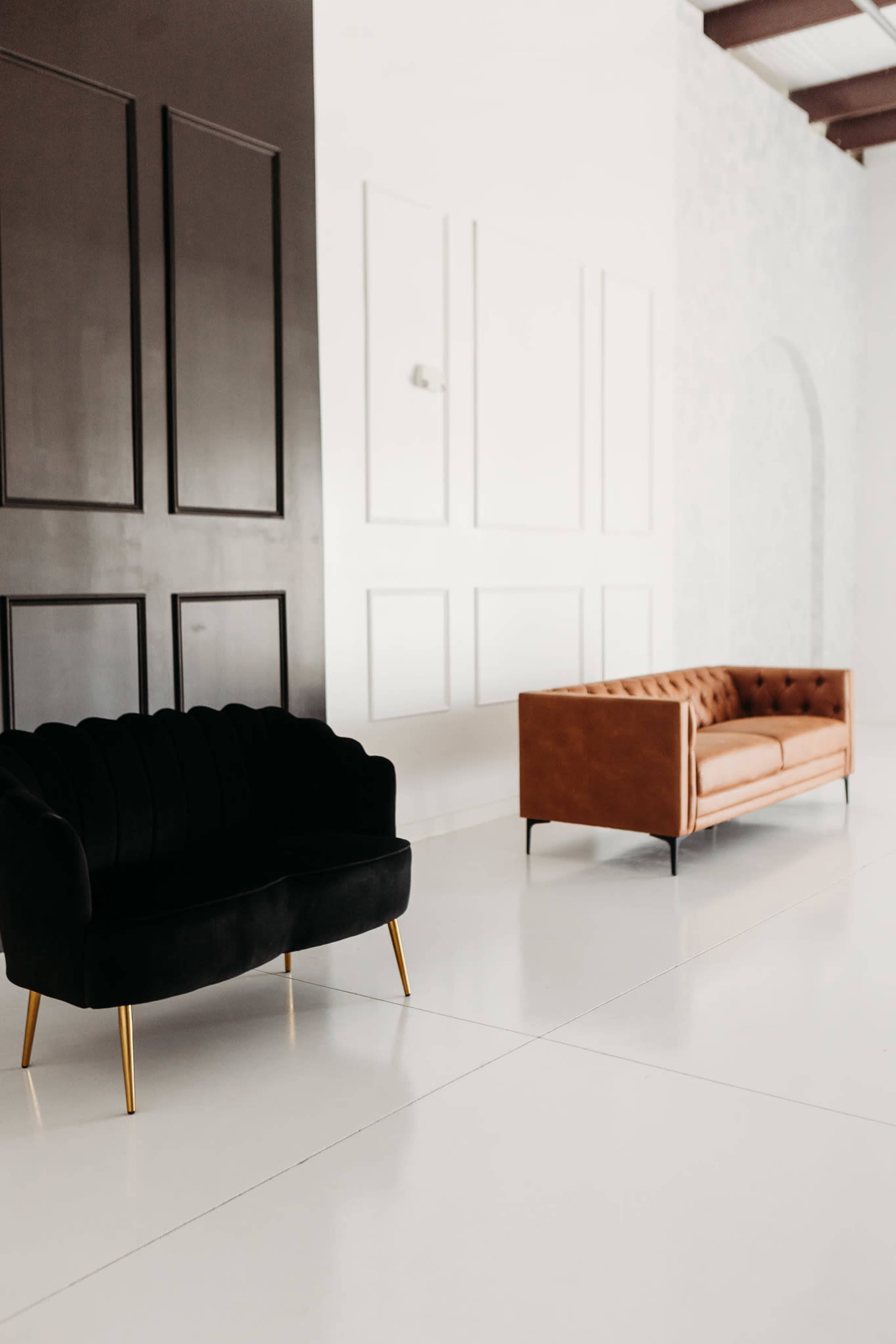 The image shows a minimalist interior with a black tufted sofa and a tan leather couch placed against a white wall.