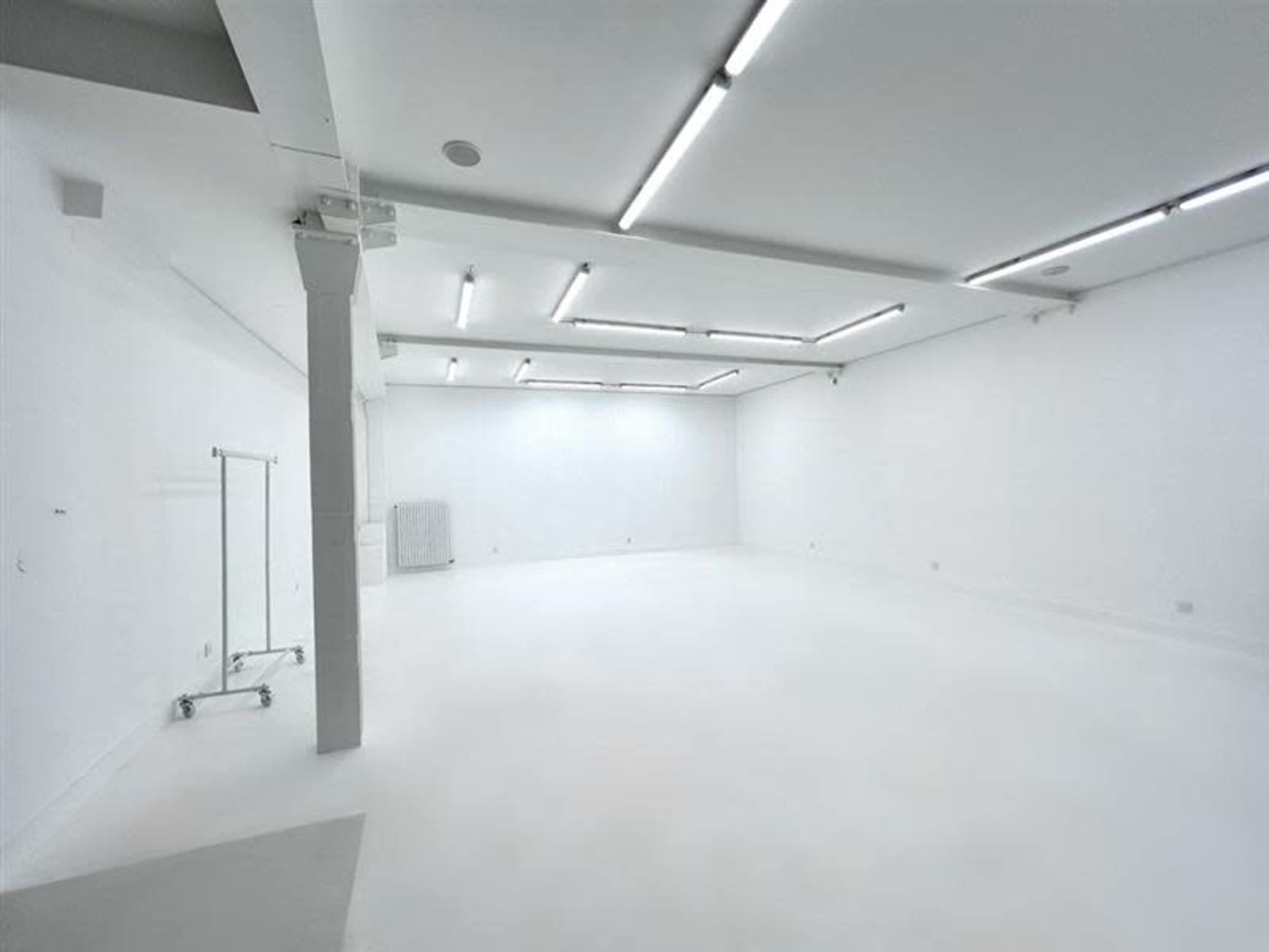 The image shows a spacious, empty room with white walls, a white floor, and fluorescent lighting.