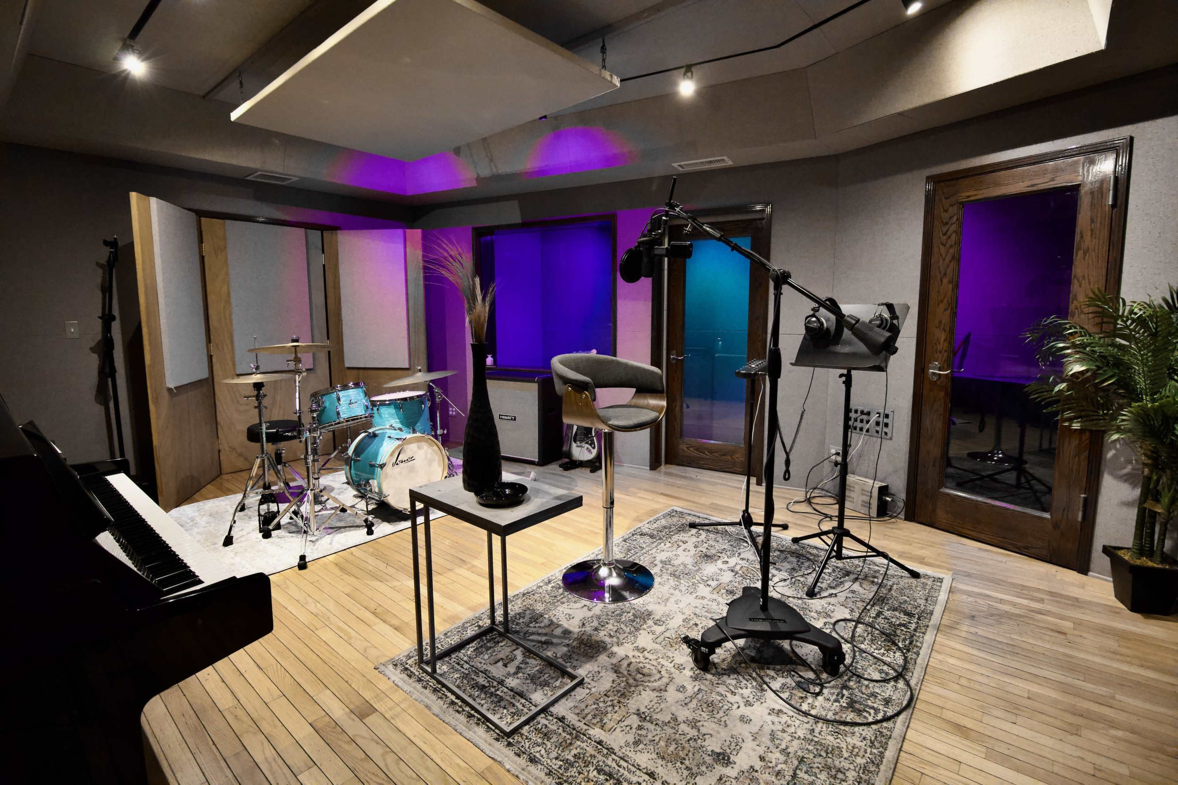 A music studio features a drum set, a piano, a microphone setup, and ambient lighting along with carpeted flooring and plants.