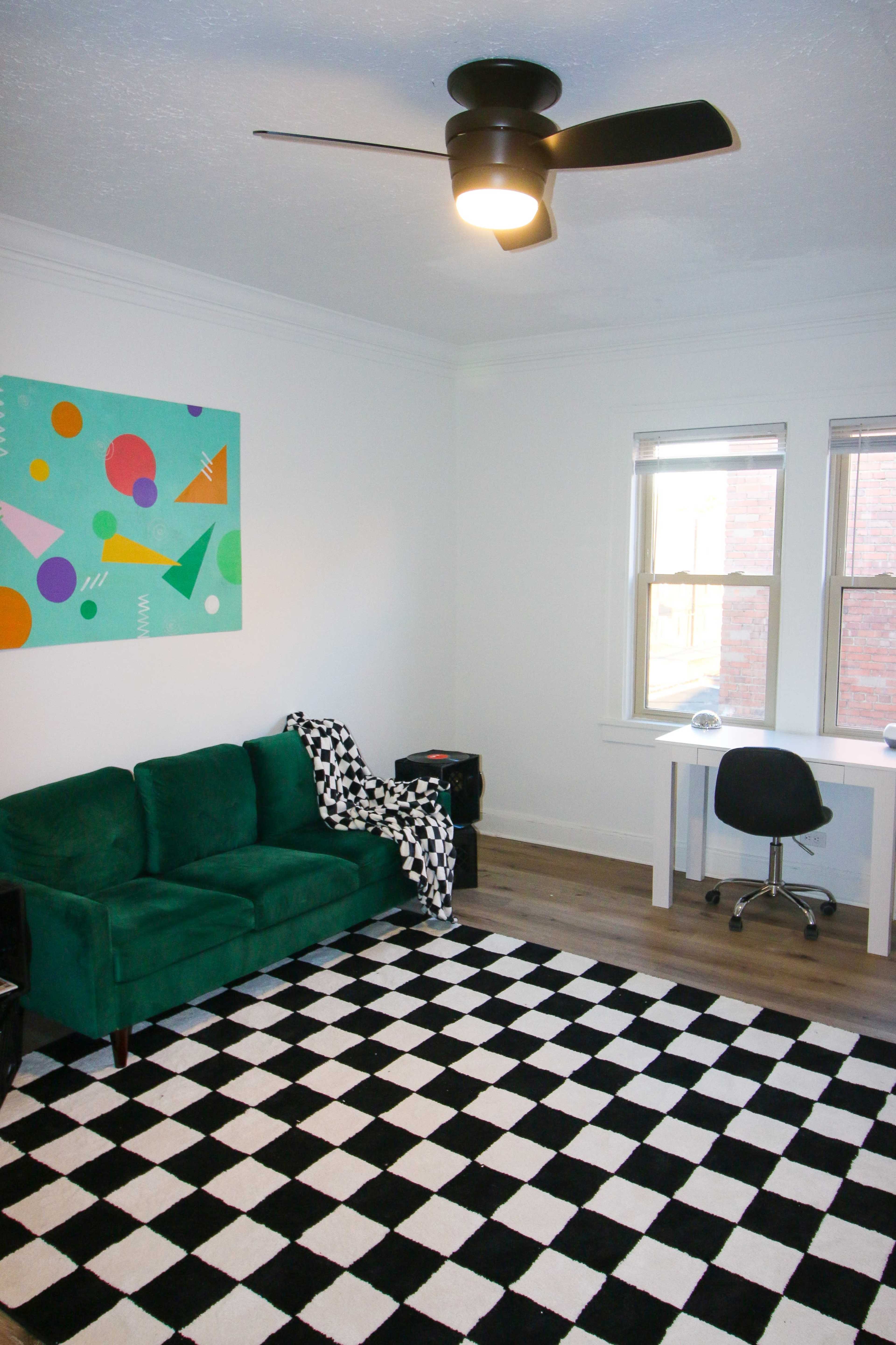 A room with a green couch, a black and white checkered rug, a desk and chair, and a colorful abstract painting on the wall.