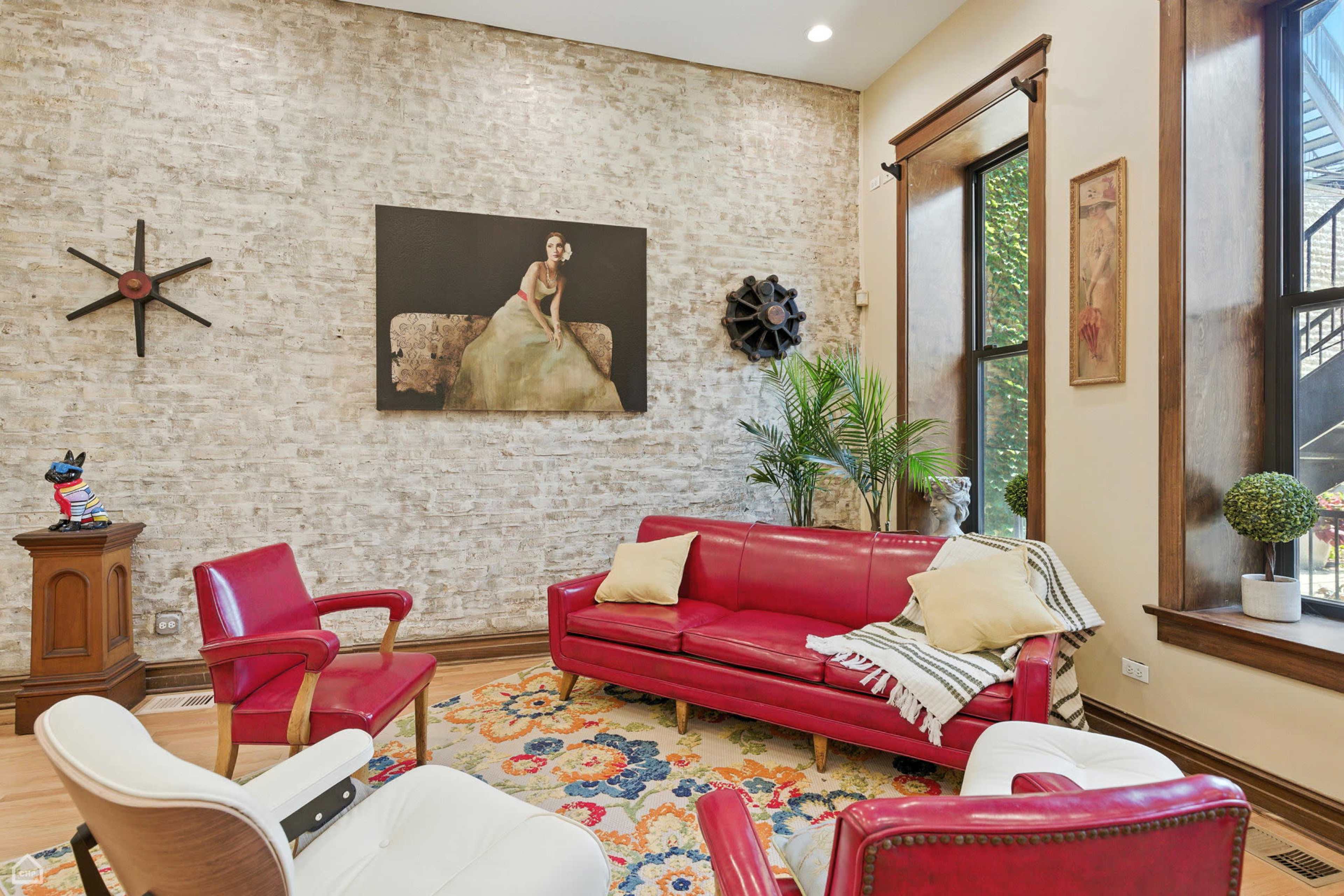 The room features a red leather couch and chairs arranged around a colorful rug, with a brick wall and large windows showcasing artwork and plants.