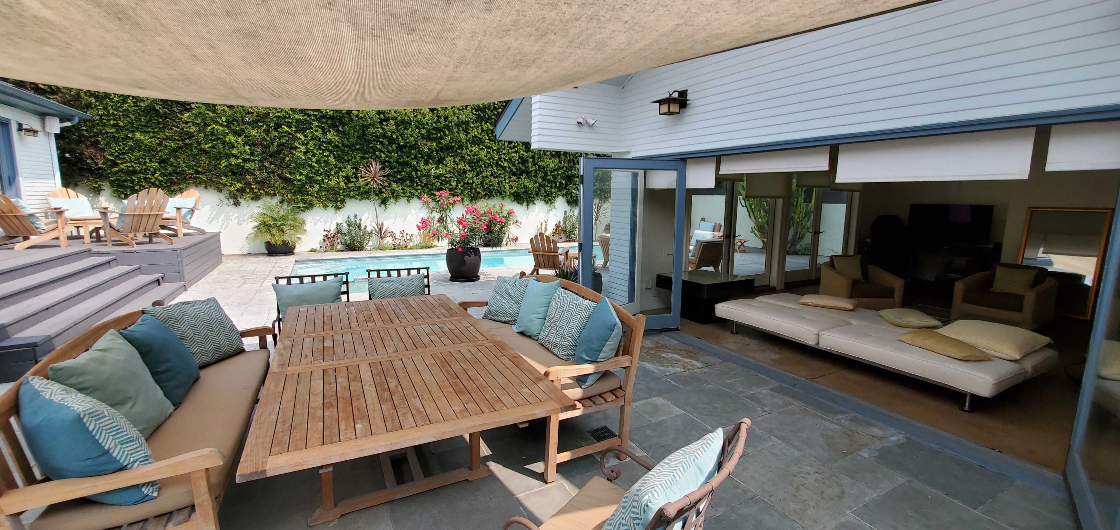 A patio area features a wooden dining table surrounded by chairs, with a pool visible in the background and a lounge area inside the house.