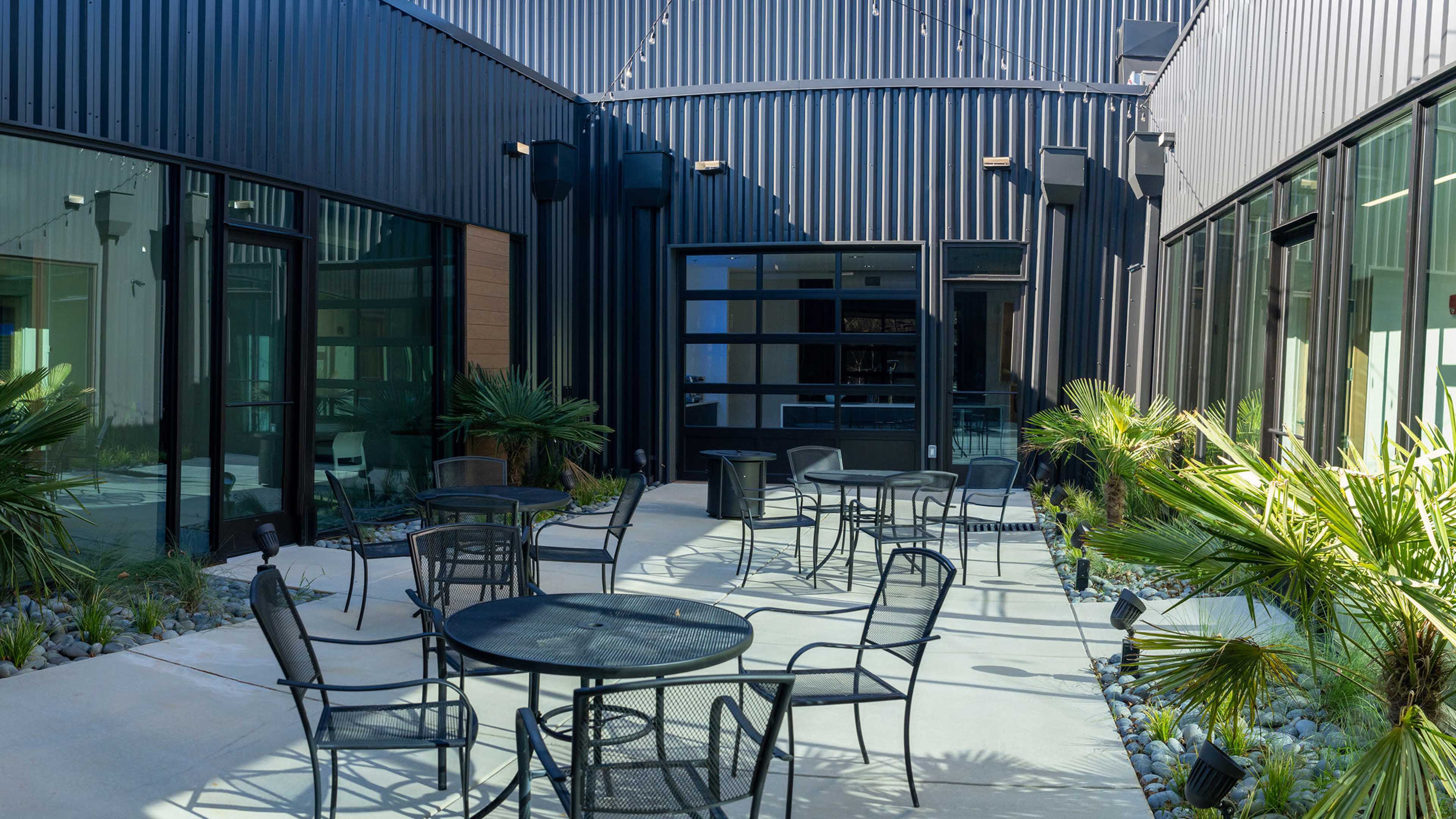 The image shows a courtyard with several black metal tables and chairs surrounded by plants and modern building facades.