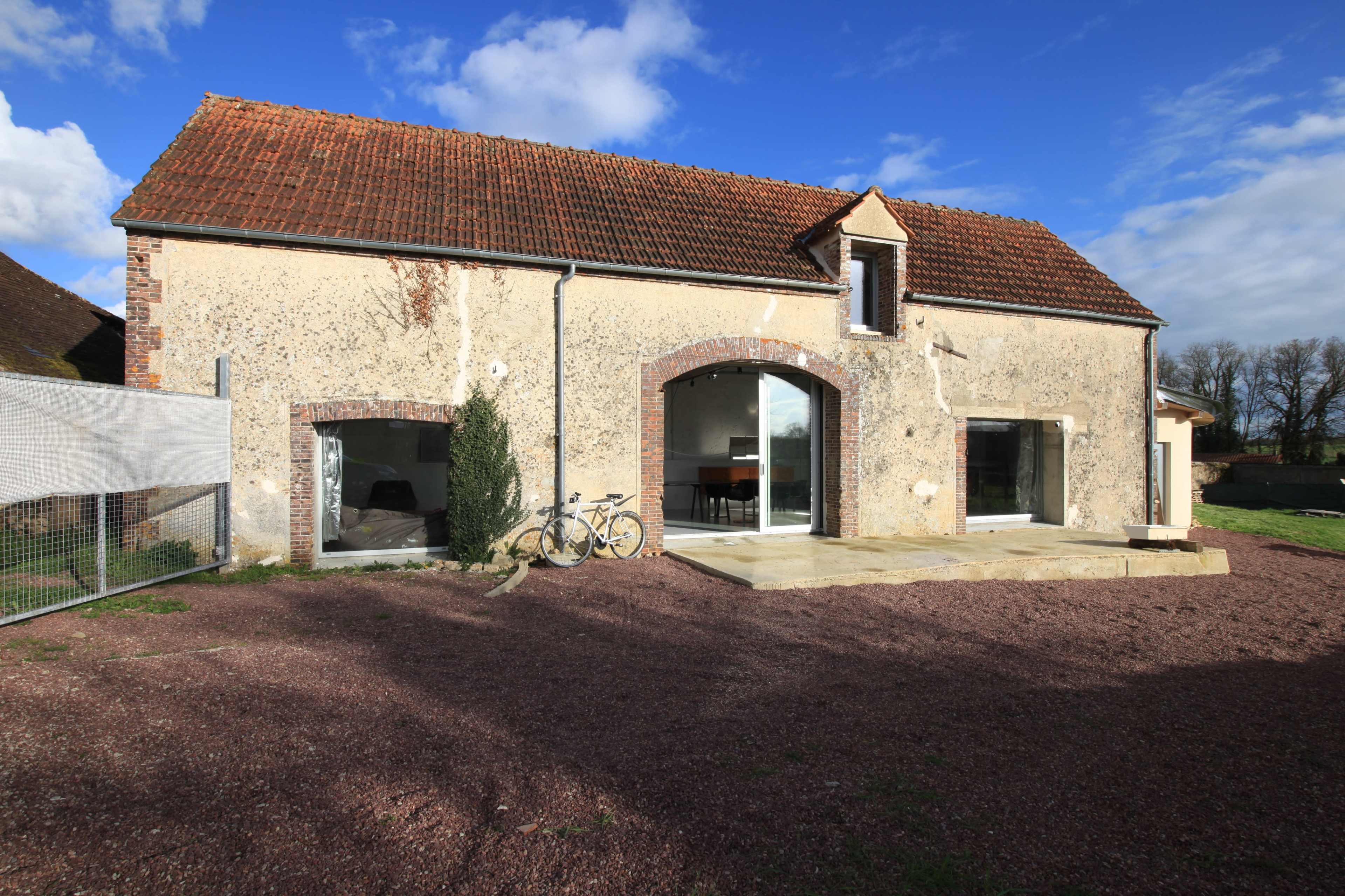 A stone house with a red-tiled roof features large windows and a small patio area, set against a blue sky with scattered clouds.