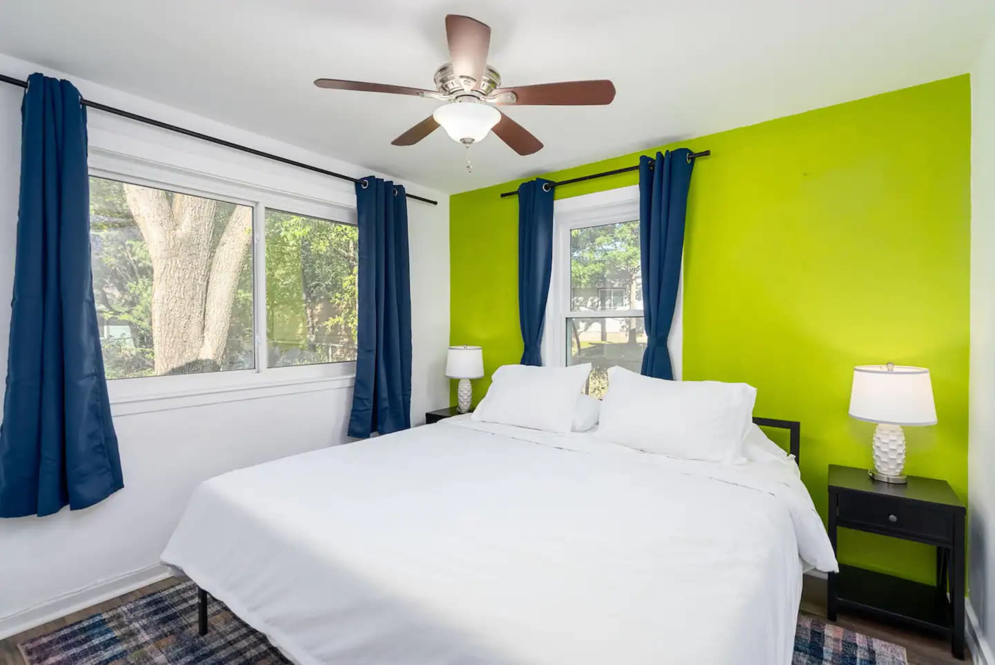 The image shows a bedroom featuring a large bed with white linens, two bedside lamps, dark blue curtains, and a bright green accent wall.