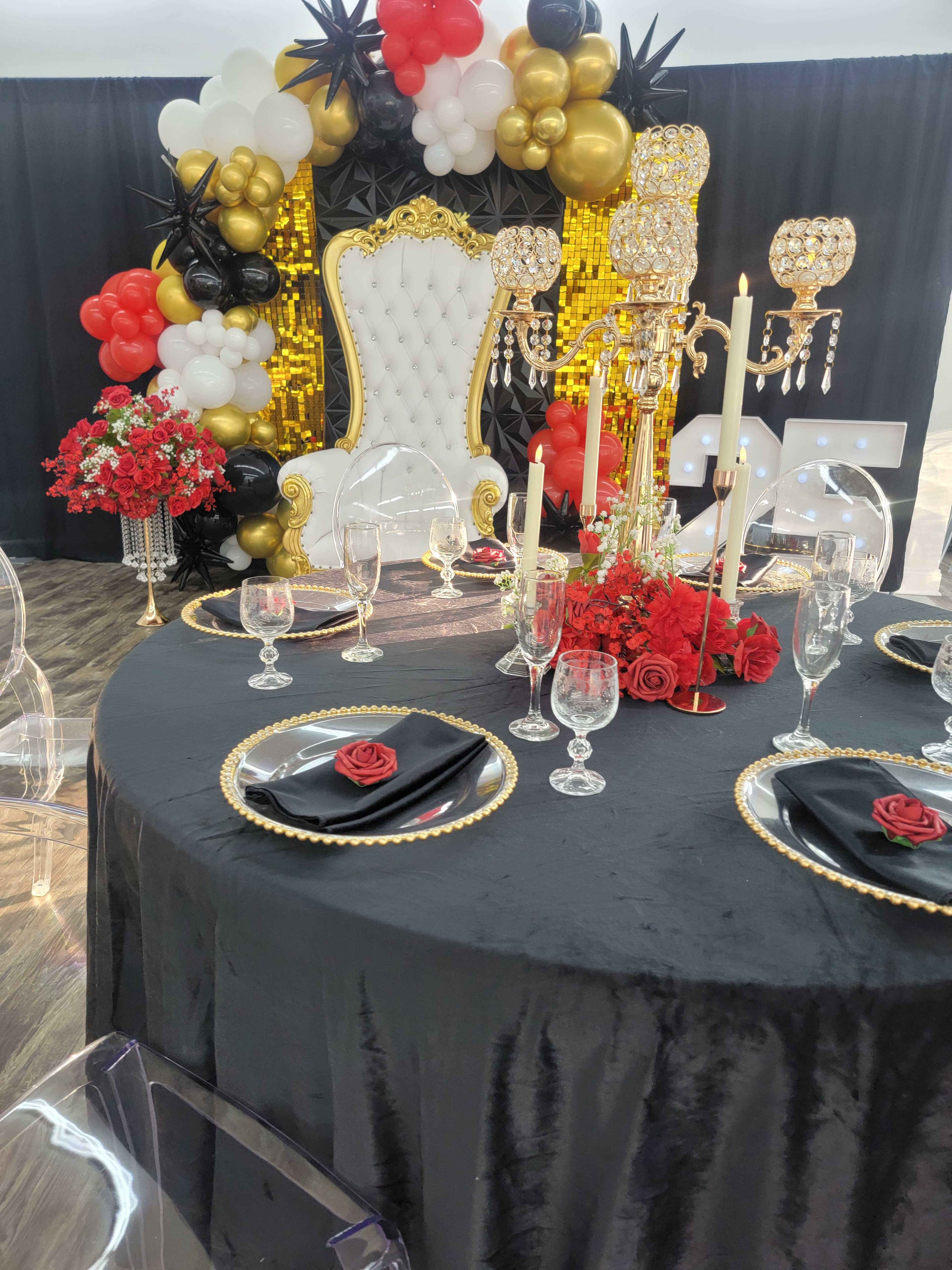 The image shows a beautifully arranged event setup featuring an elegantly decorated table with black and gold accents, a white throne chair, and a backdrop of balloons in black, gold, and red.