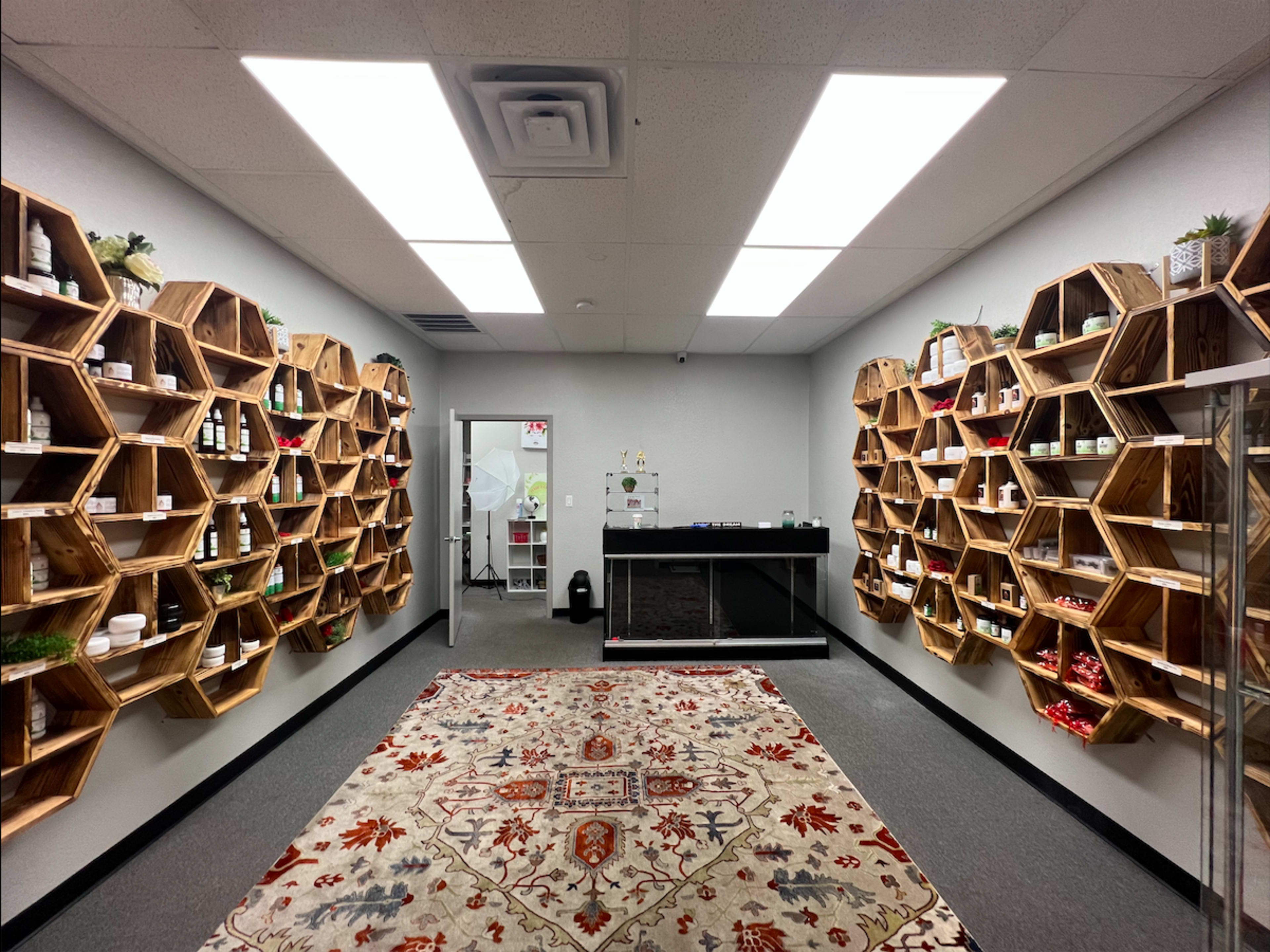 The image shows a spacious retail room with wooden hexagonal shelves displaying various products along the walls, a reception counter in the center, and a patterned rug on the floor.