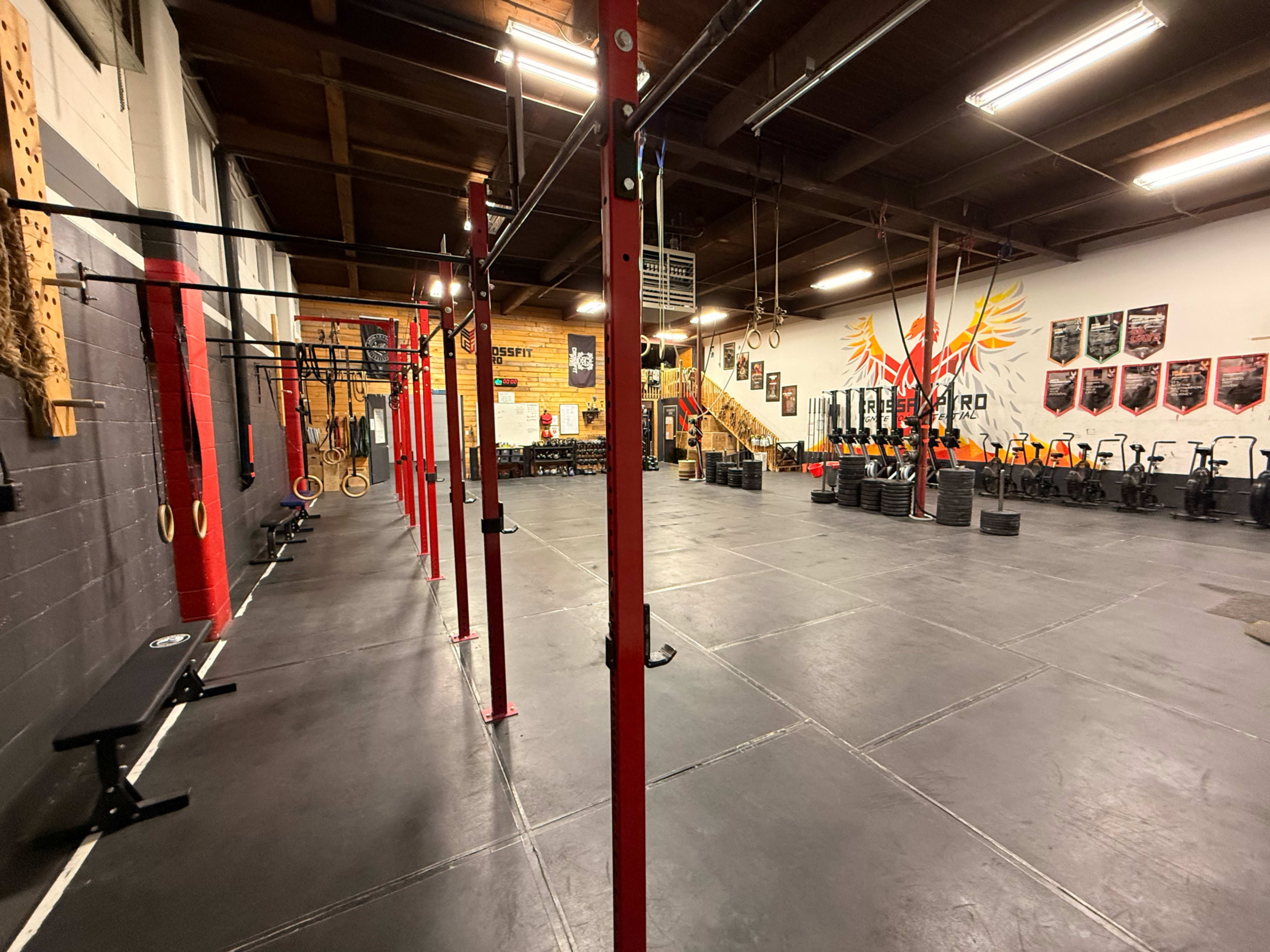 The image shows an empty gym space with black rubber flooring, red pull-up bars, and various fitness equipment arranged along the walls.