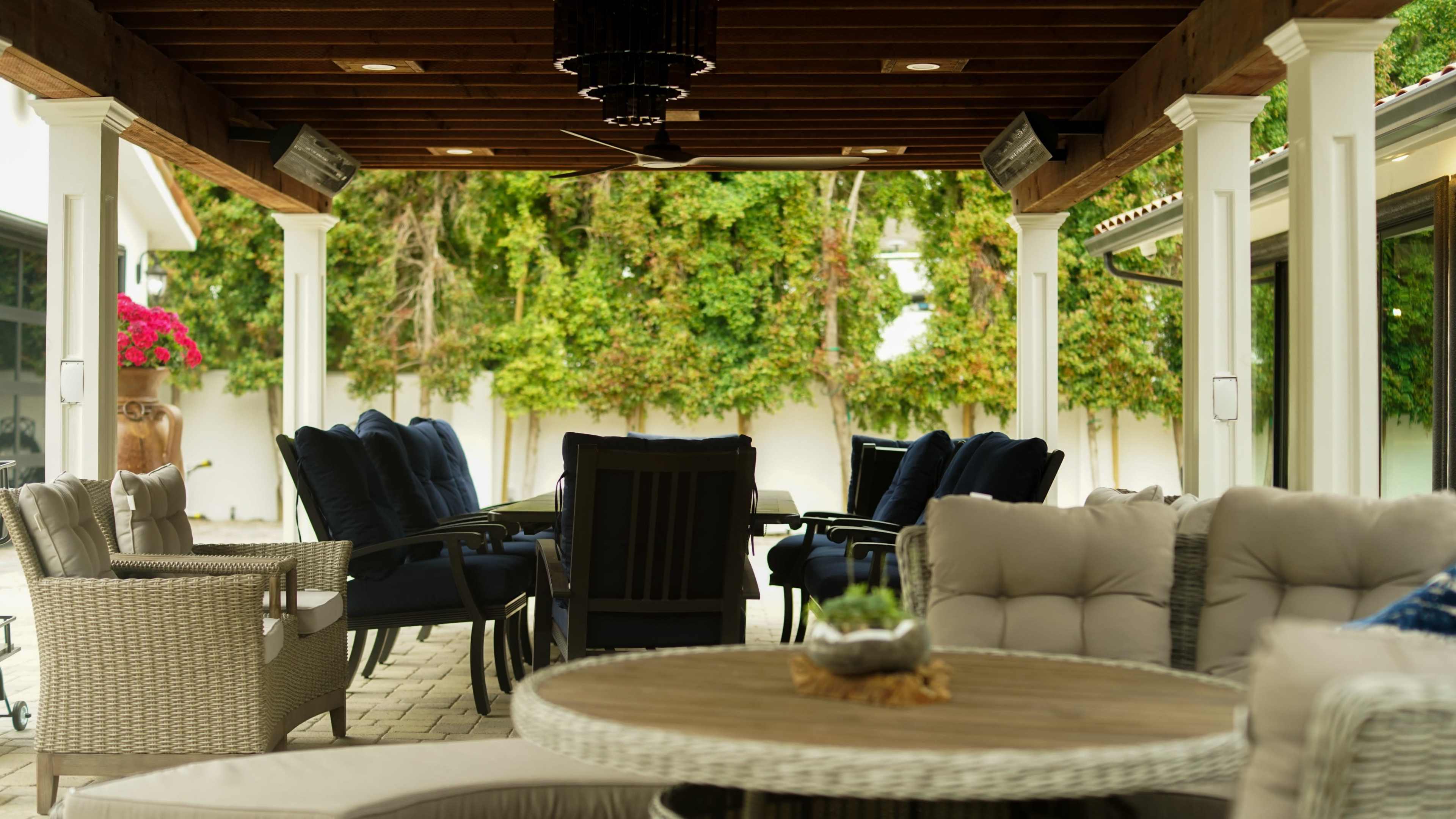 A covered outdoor patio features several seating arrangements with a round table at the center, surrounded by lush green foliage and decorative plants.