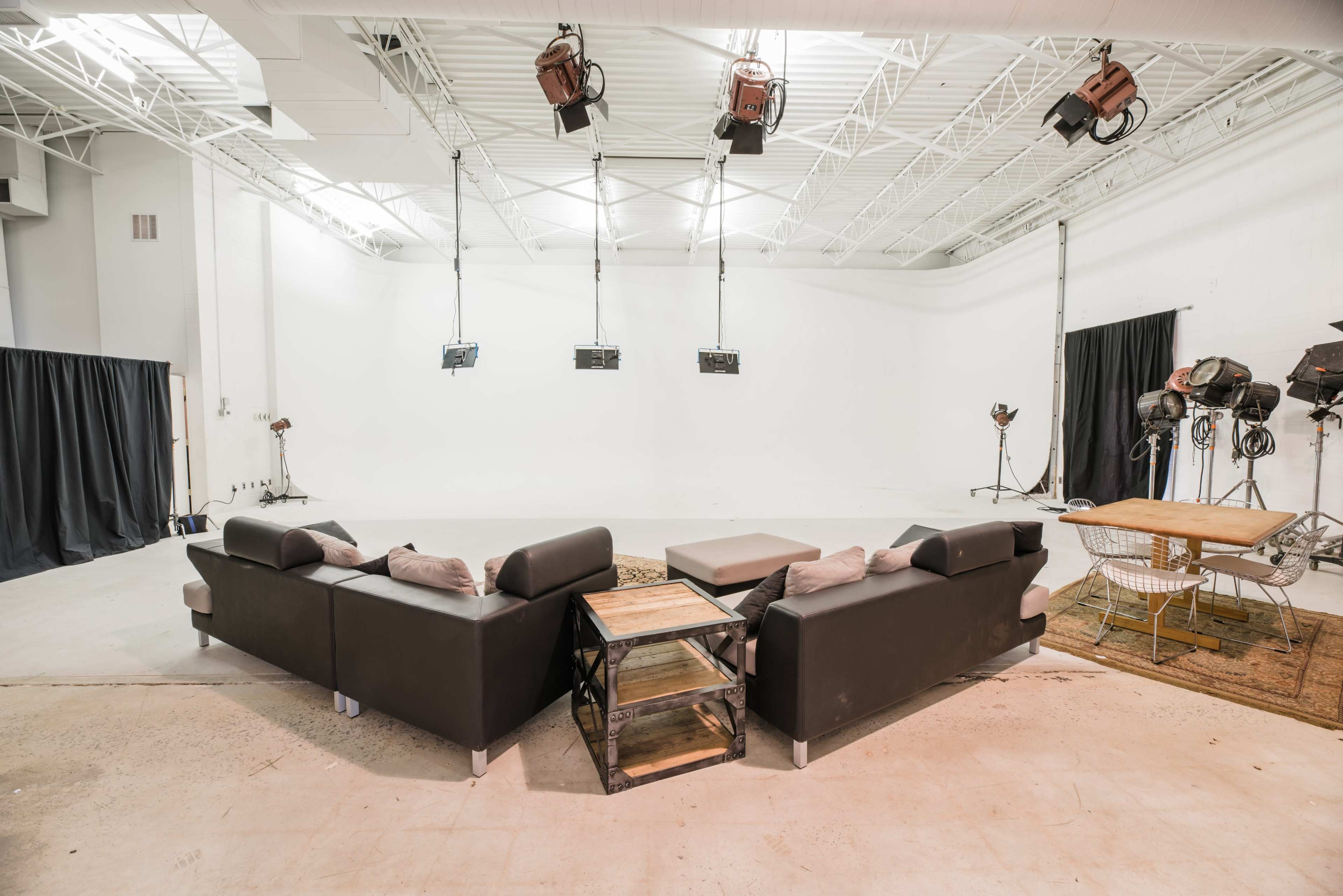 A spacious studio features a black couch arrangement, a wooden coffee table, and various lighting equipment positioned against a white backdrop.
