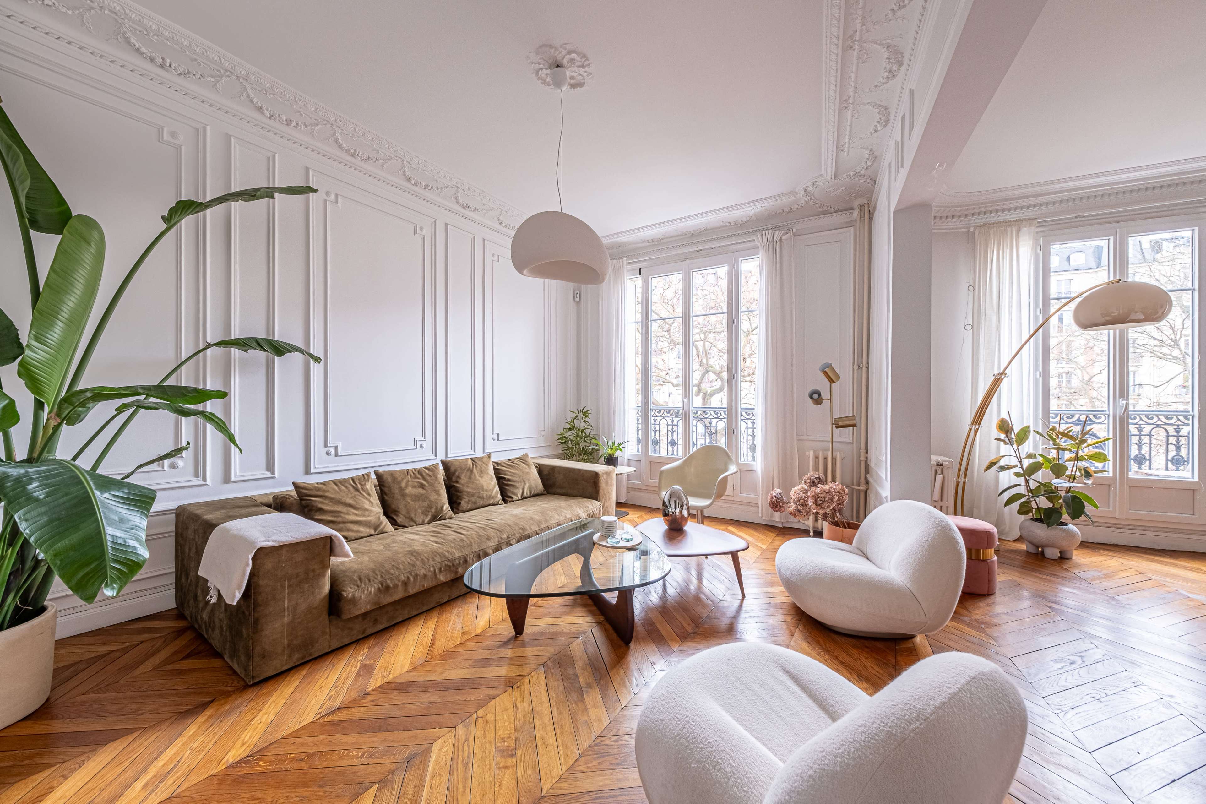 A spacious living room features a brown sofa, a glass coffee table, and two white chairs, surrounded by large windows and decorative moulding on the walls.
