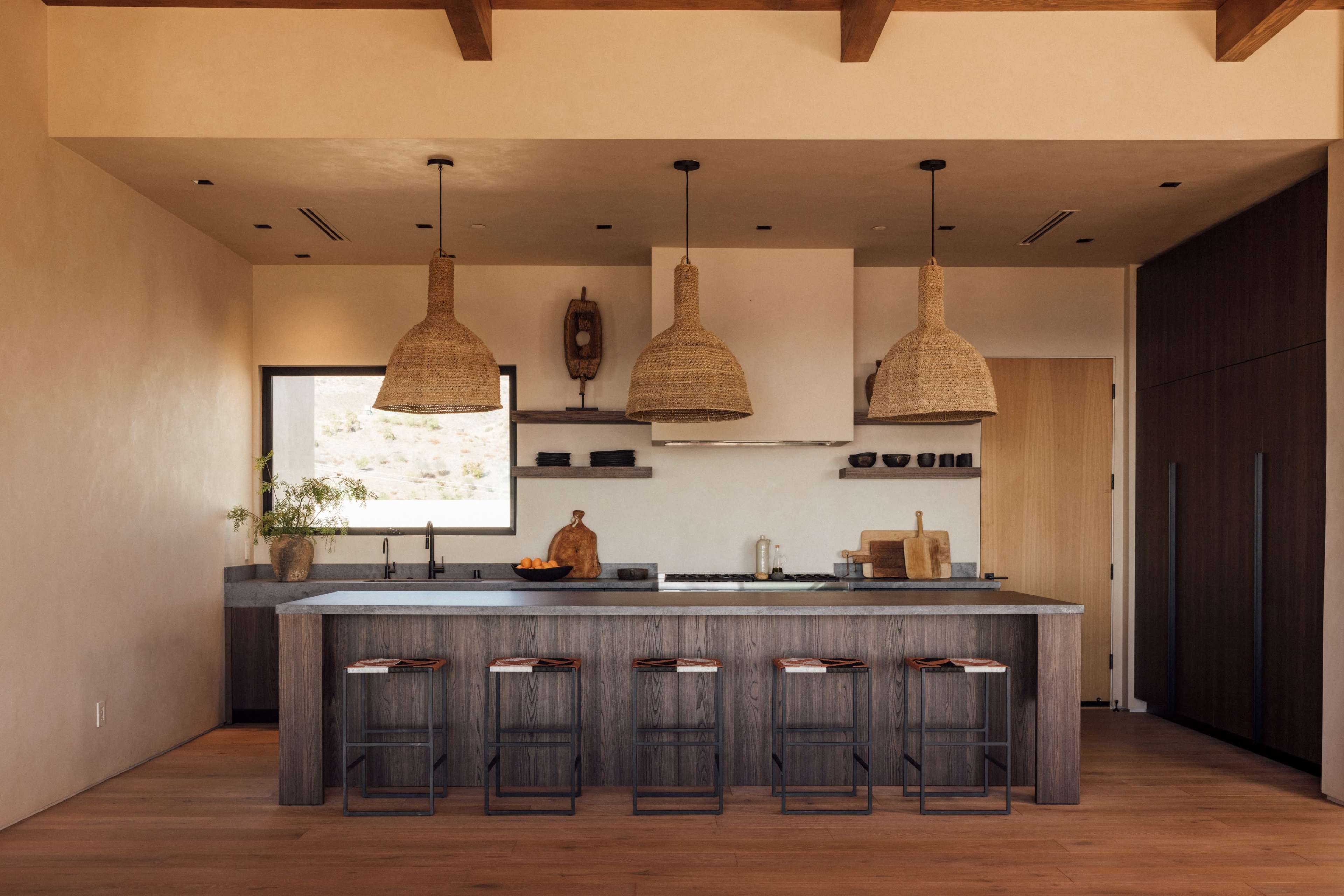 A modern kitchen features a large island with bar stools, wooden cabinetry, and woven pendant lights.