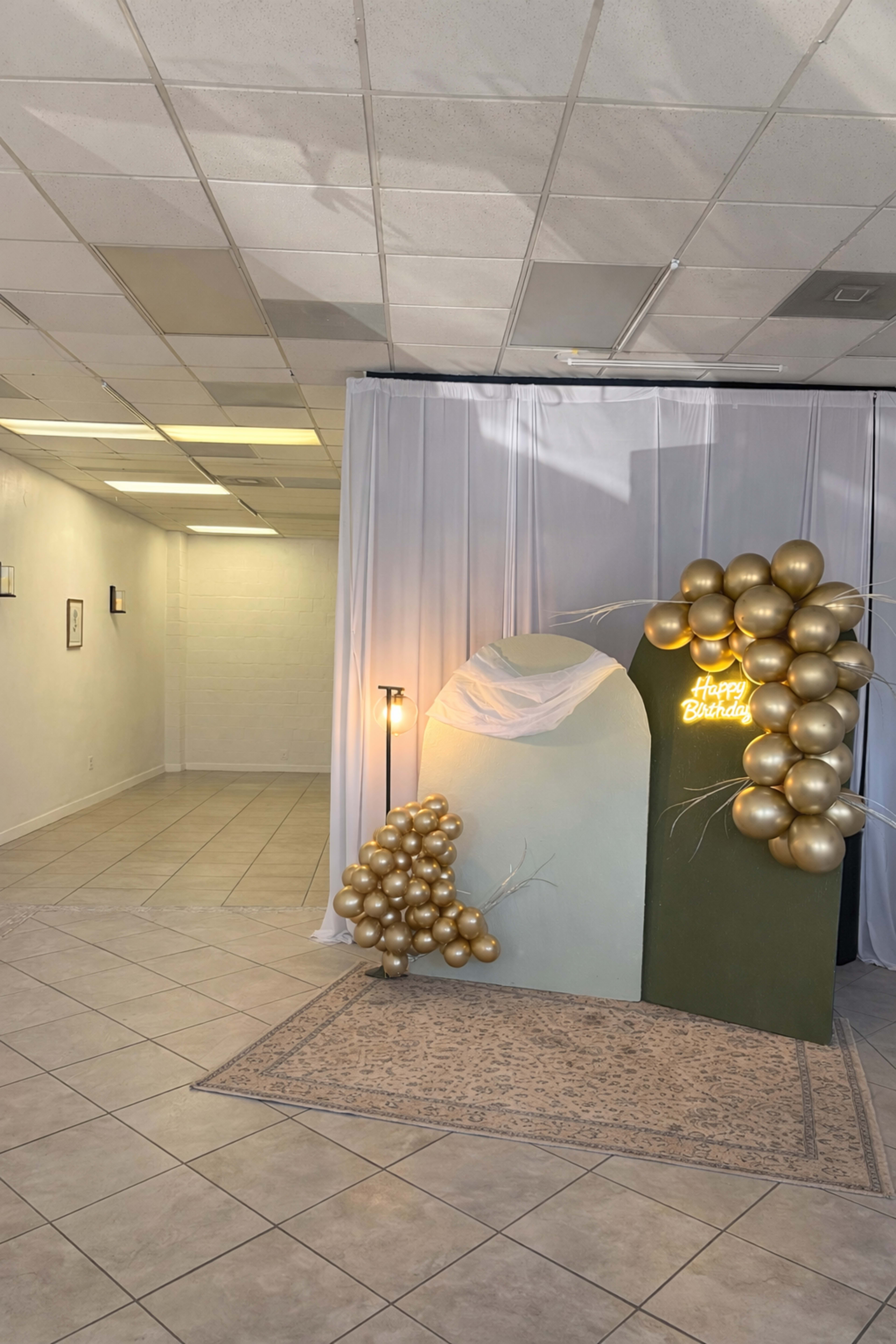 The image shows a decorated corner of a room with a birthday-themed backdrop featuring balloons and a sign that reads "Happy Birthday," along with a rug on the floor.