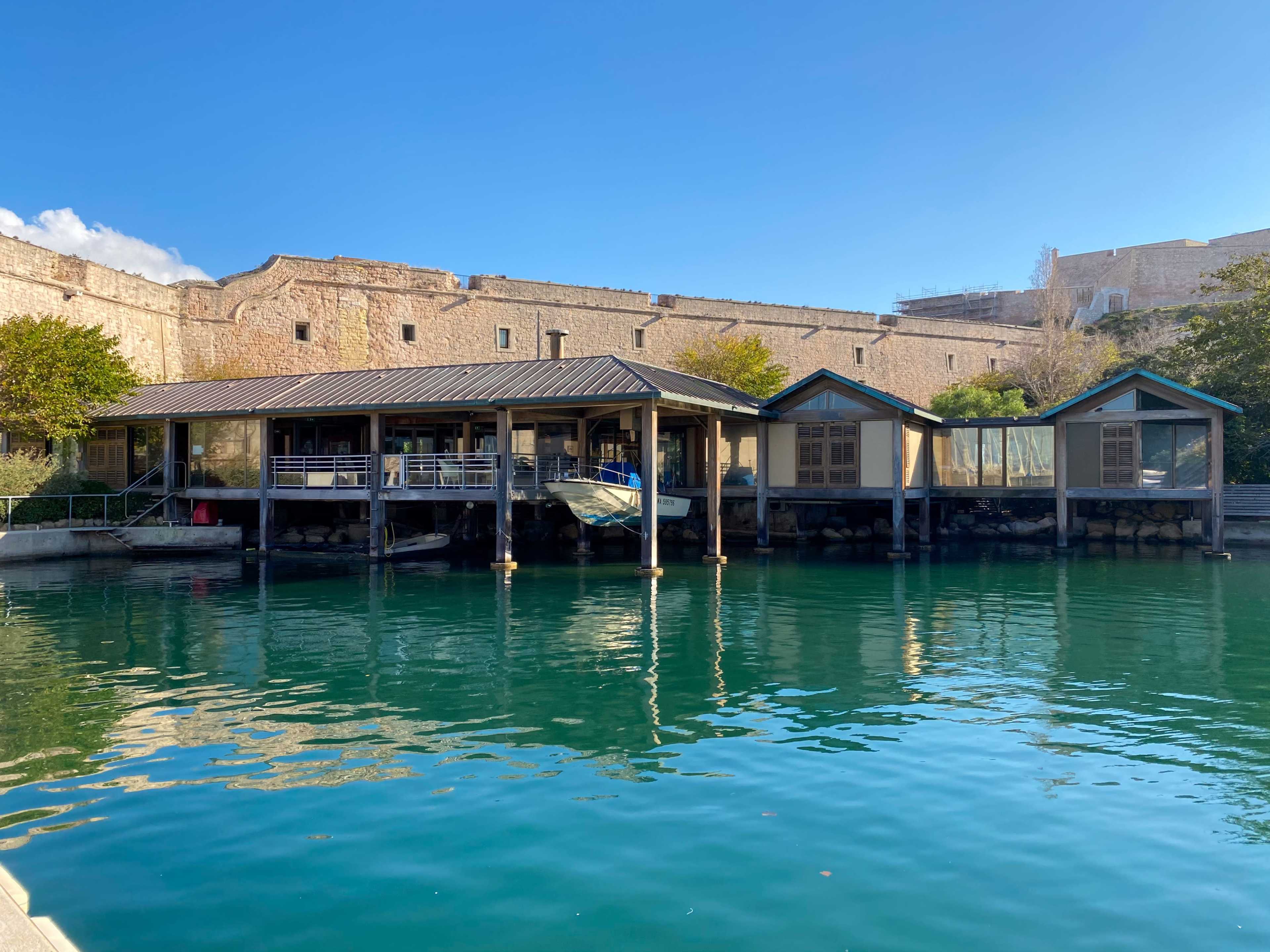 A wooden structure with a sloped roof extends over water, positioned against a stone wall in the background.