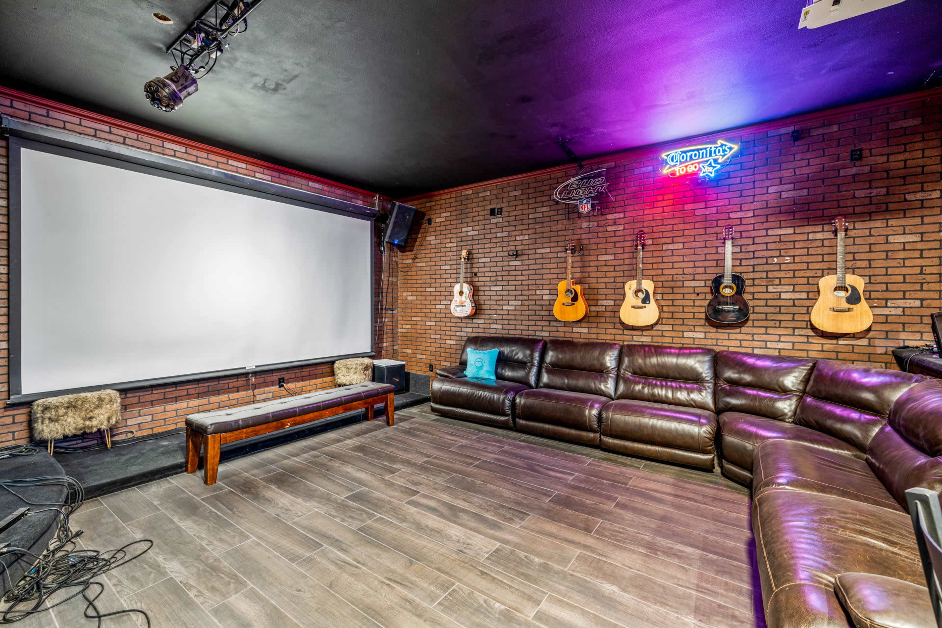 The image shows a lounge area with a large screen at one end, surrounded by a brick wall and several acoustic guitars hanging above a leather sectional sofa.