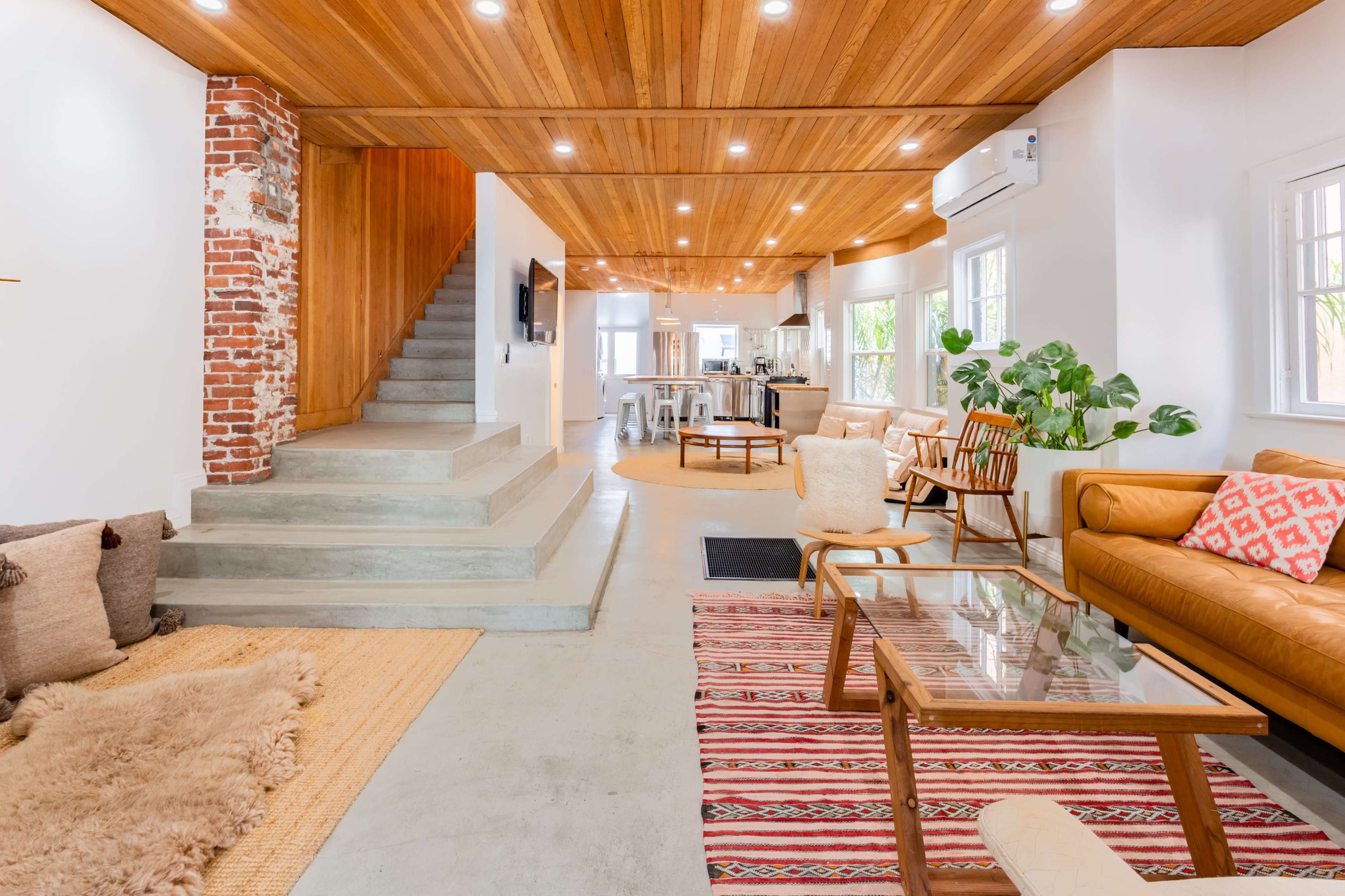 A modern living room with wooden ceilings, a staircase leading to an upper level, and a mix of seating areas, including a leather sofa and a glass coffee table.