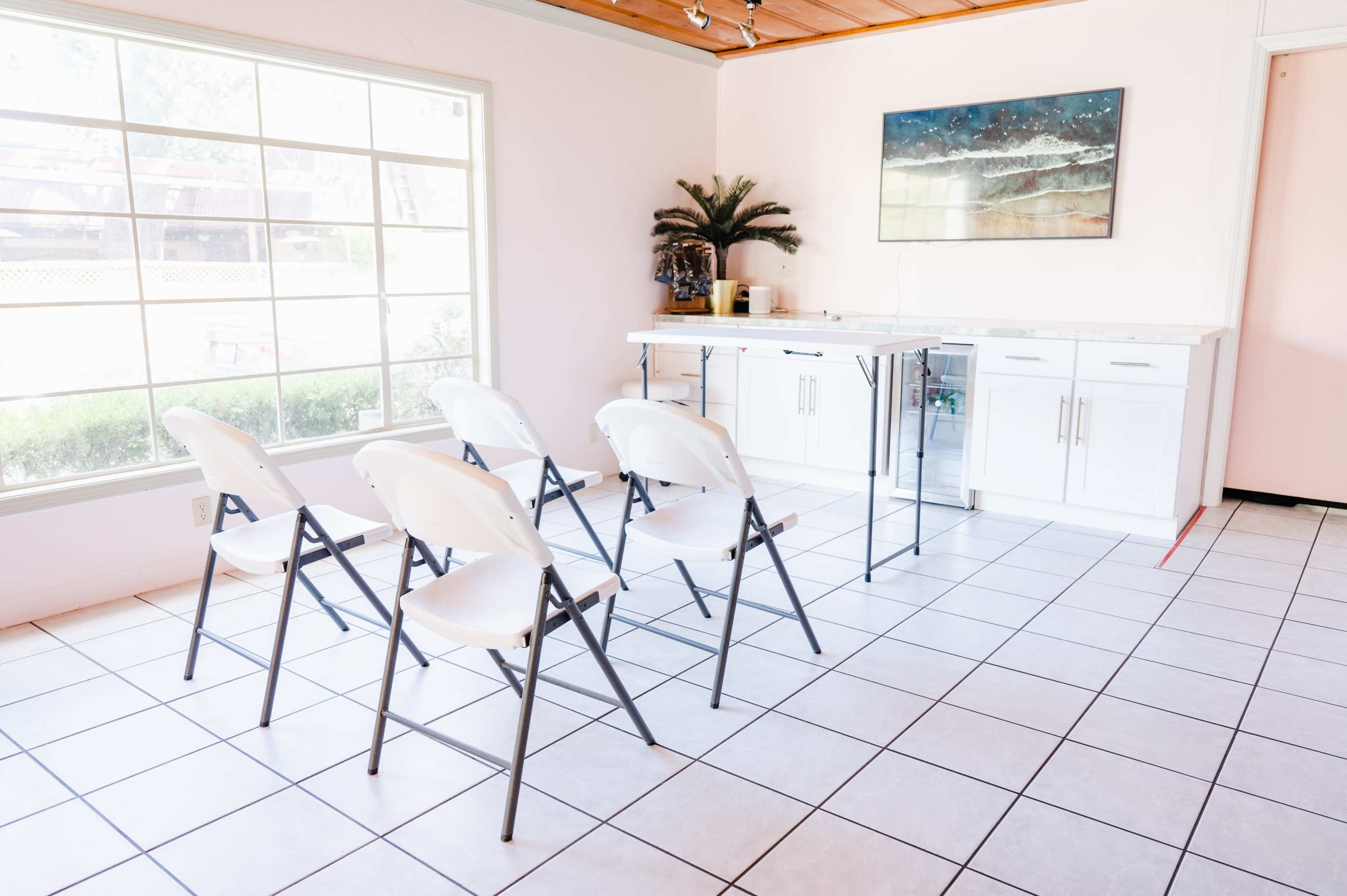 A bright room features a row of six folding chairs facing a small counter with a modern painting on the wall.