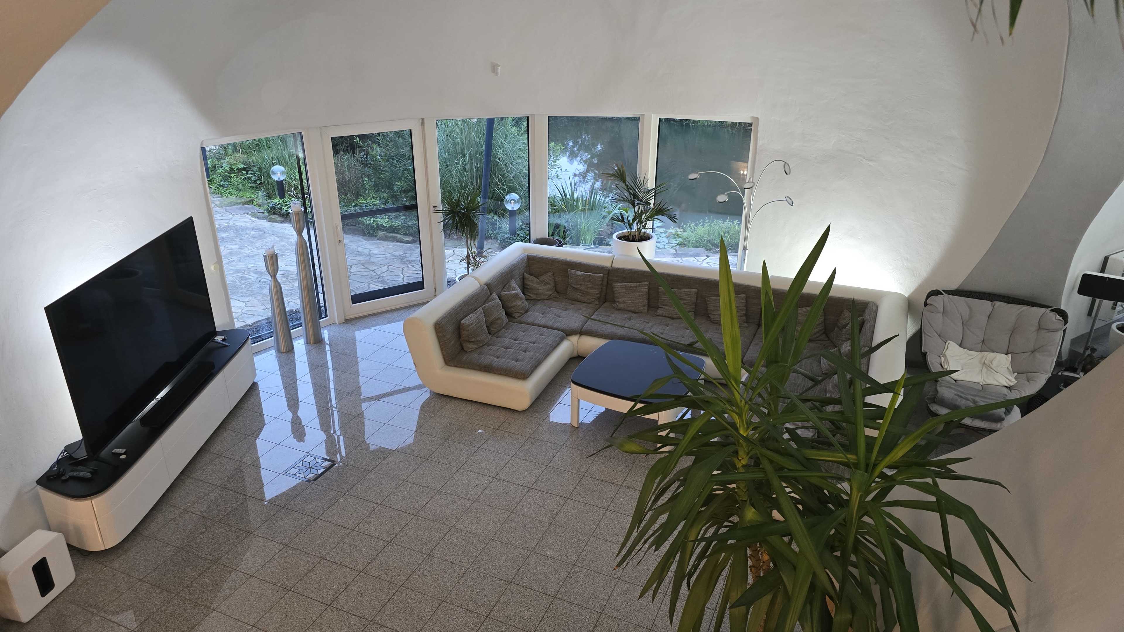 The image shows a spacious living area with a modern sectional sofa, a flat-screen TV, large windows providing a view of greenery, and a polished tile floor.