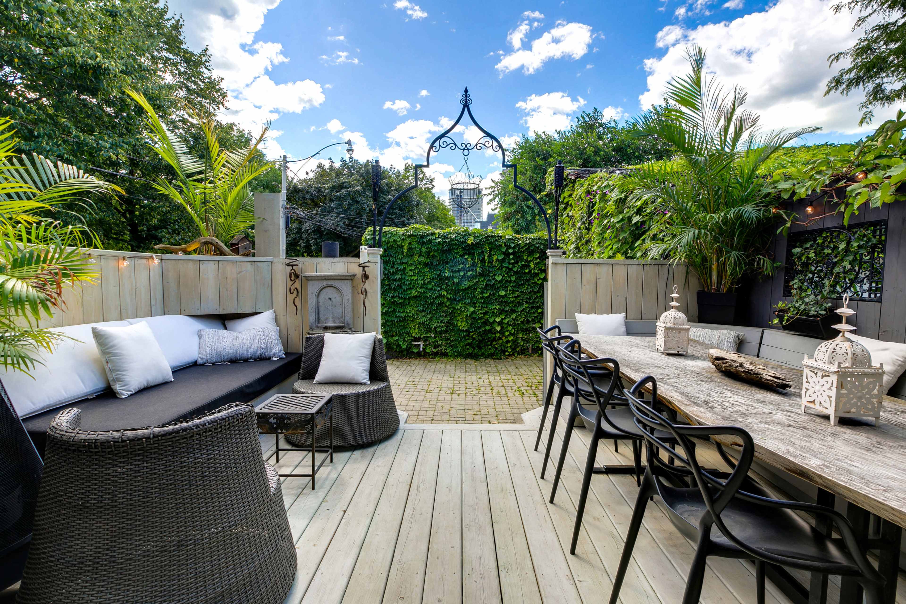 A landscaped outdoor deck area featuring a seating arrangement with a black couch, a dining table, and lush greenery surrounding the space.