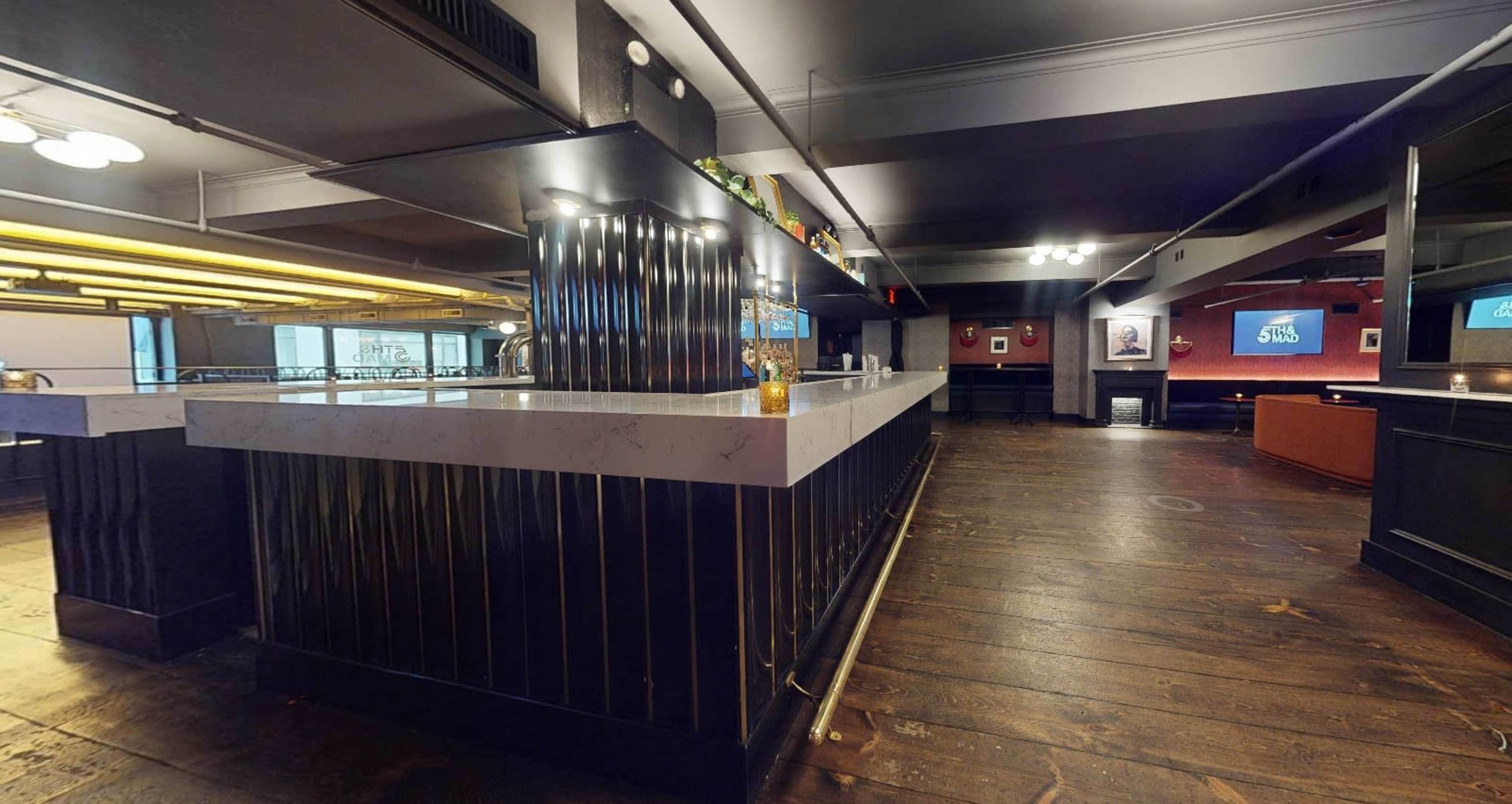 The image shows a spacious bar area with a sleek counter, wooden flooring, and multiple screens displaying content.