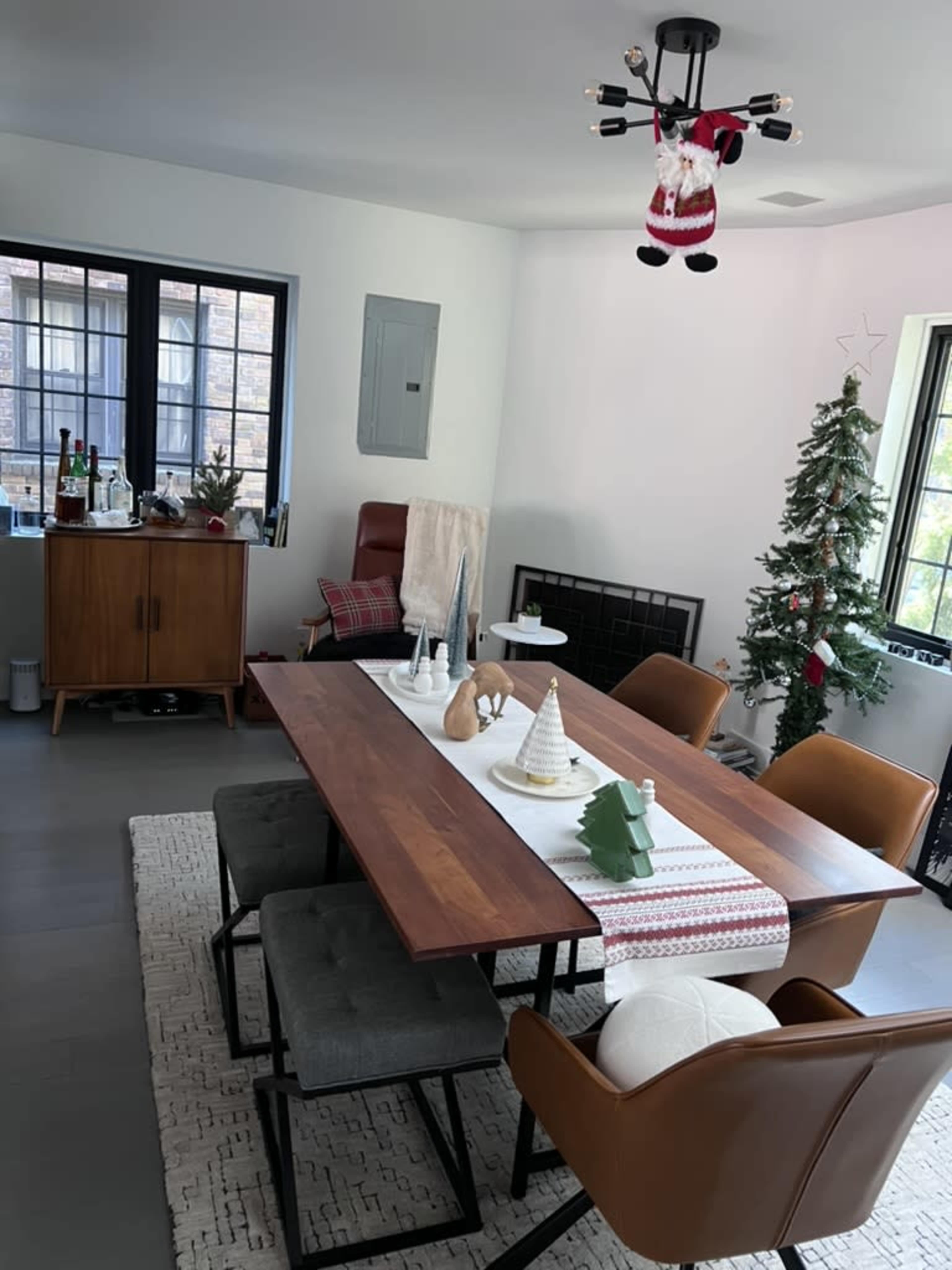 A dining room features a wooden table surrounded by chairs, decorated with a holiday table runner, while a Christmas tree stands in the corner.