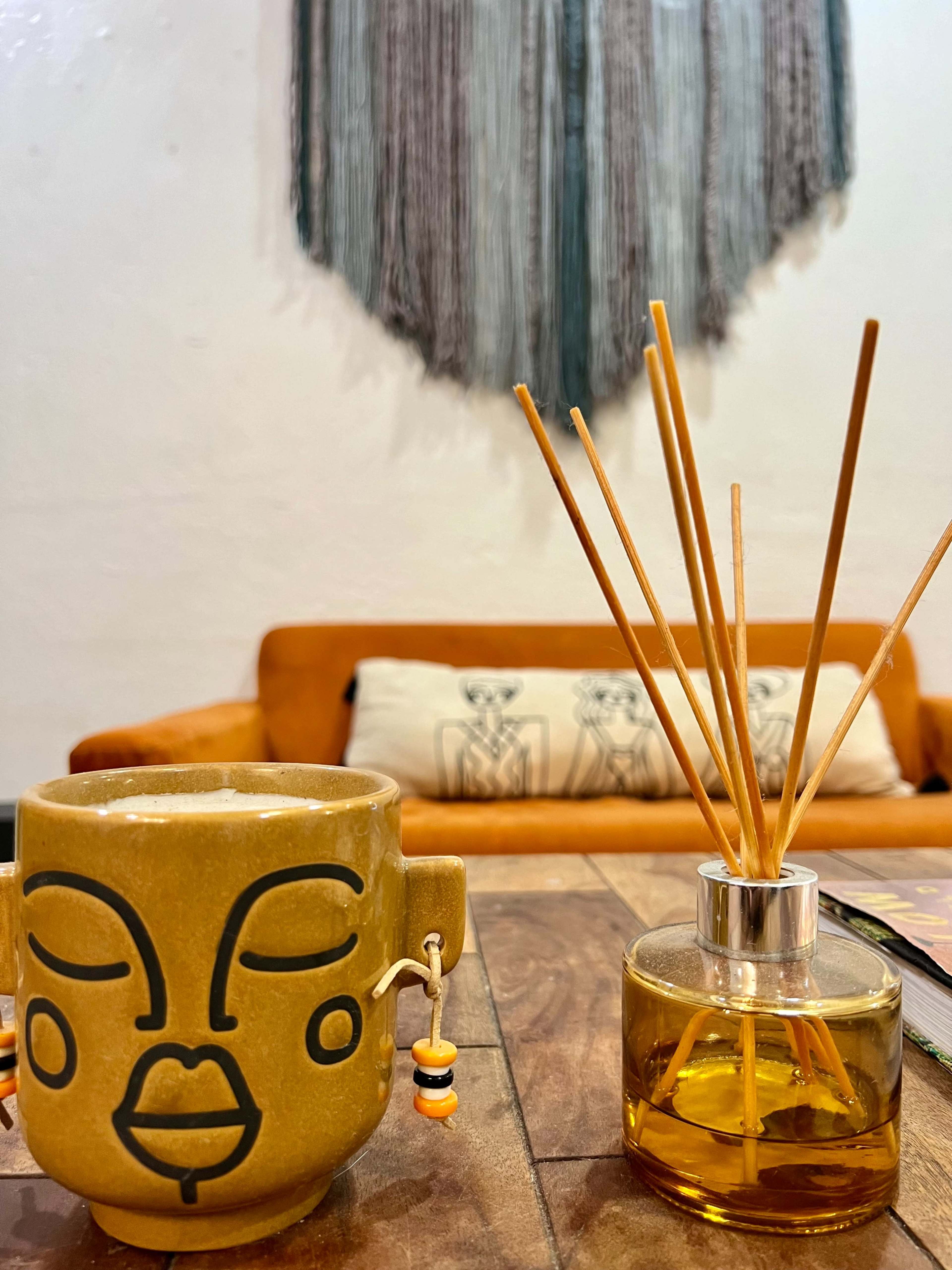 A yellow mug with a face design sits next to a diffuser with reeds on a wooden table, in front of a brown couch and a decorative wall hanging.
