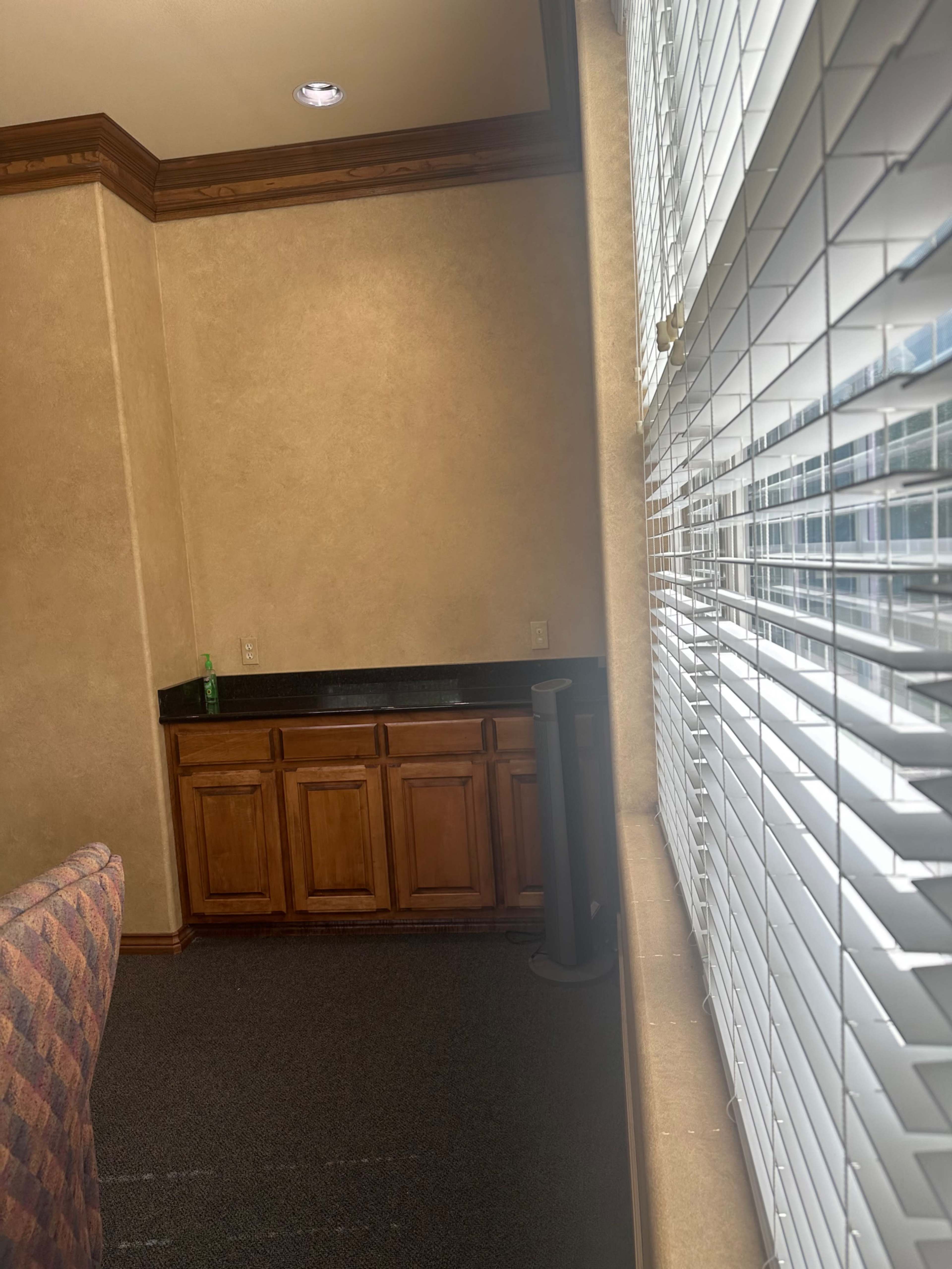 The image shows a corner of a room with a countertop, wooden cabinets, and a window covered by blinds.