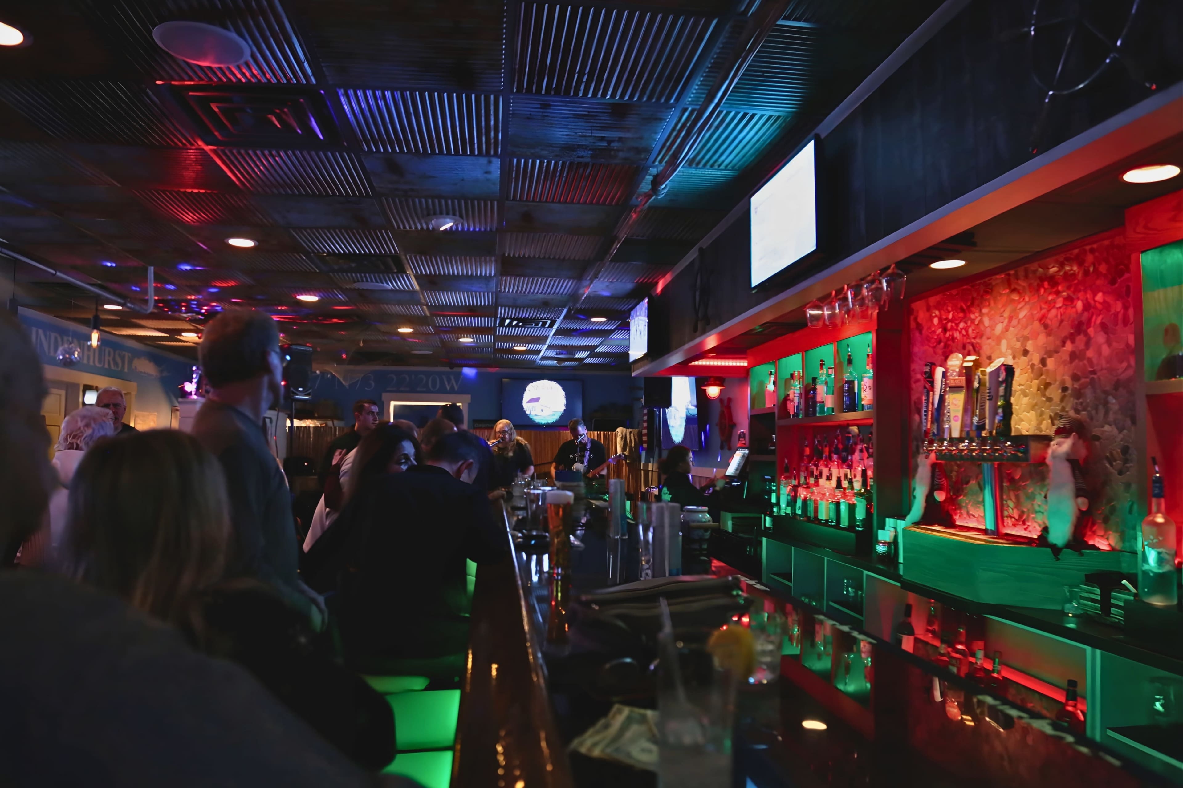The image shows a lively bar with colorful lighting, patrons seated at the counter, and various drinks displayed on the shelves behind the bar.