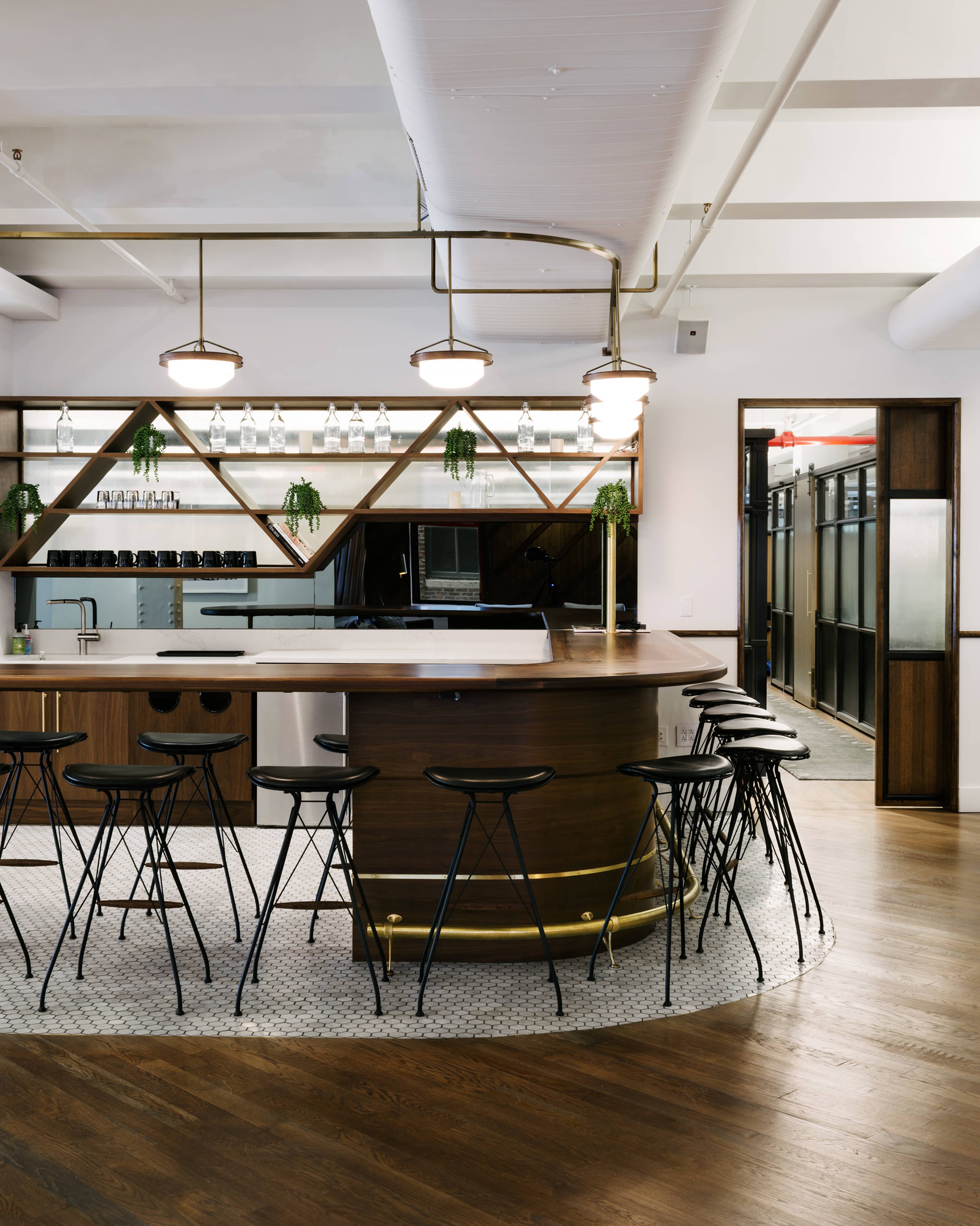 The image shows a modern bar area with a curved wooden counter, black stools, and decorative shelving featuring glassware and greenery.