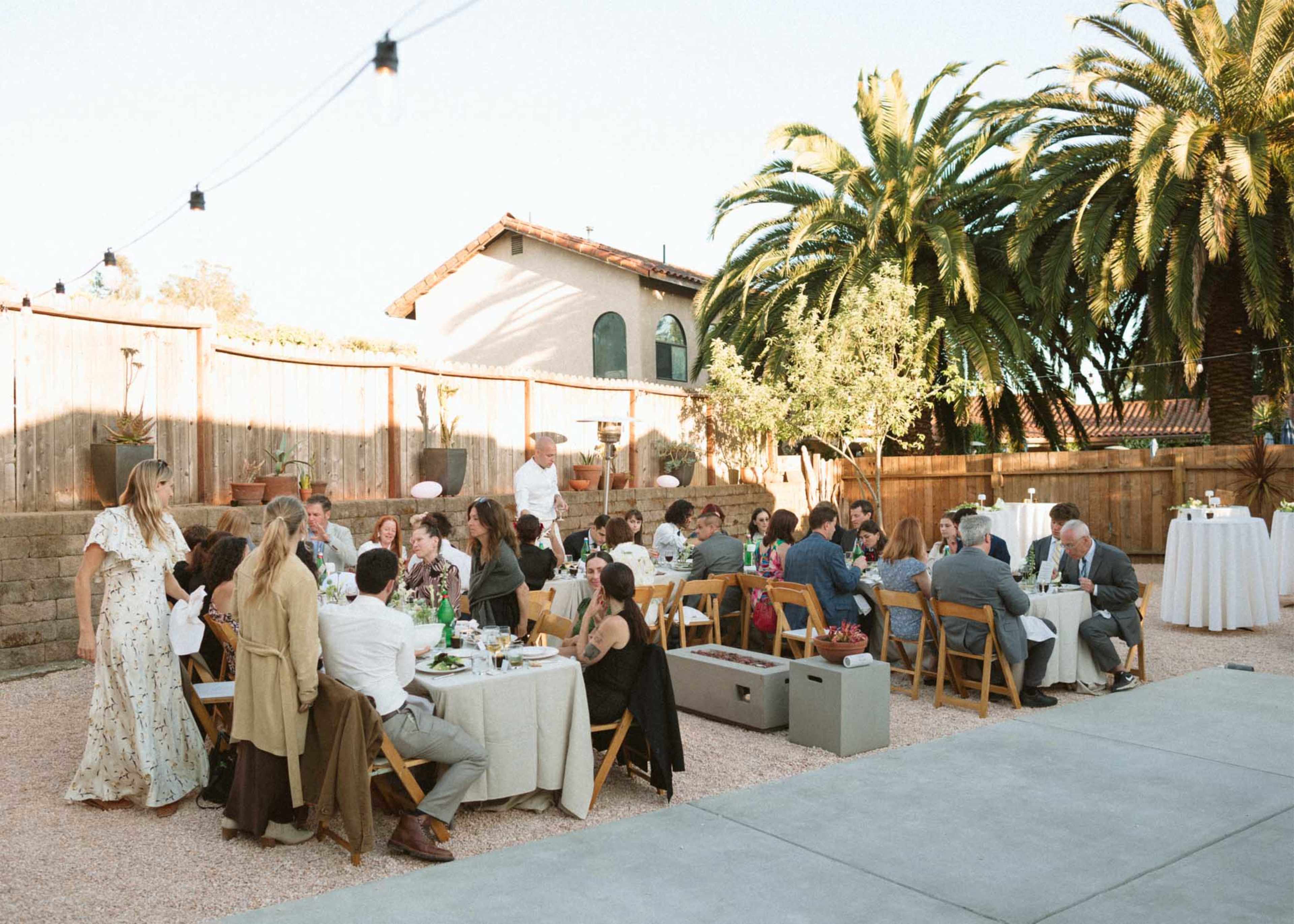 A group of guests is seated at tables outdoors, surrounded by palm trees and a wooden fence, enjoying a gathering in a well-lit setting.