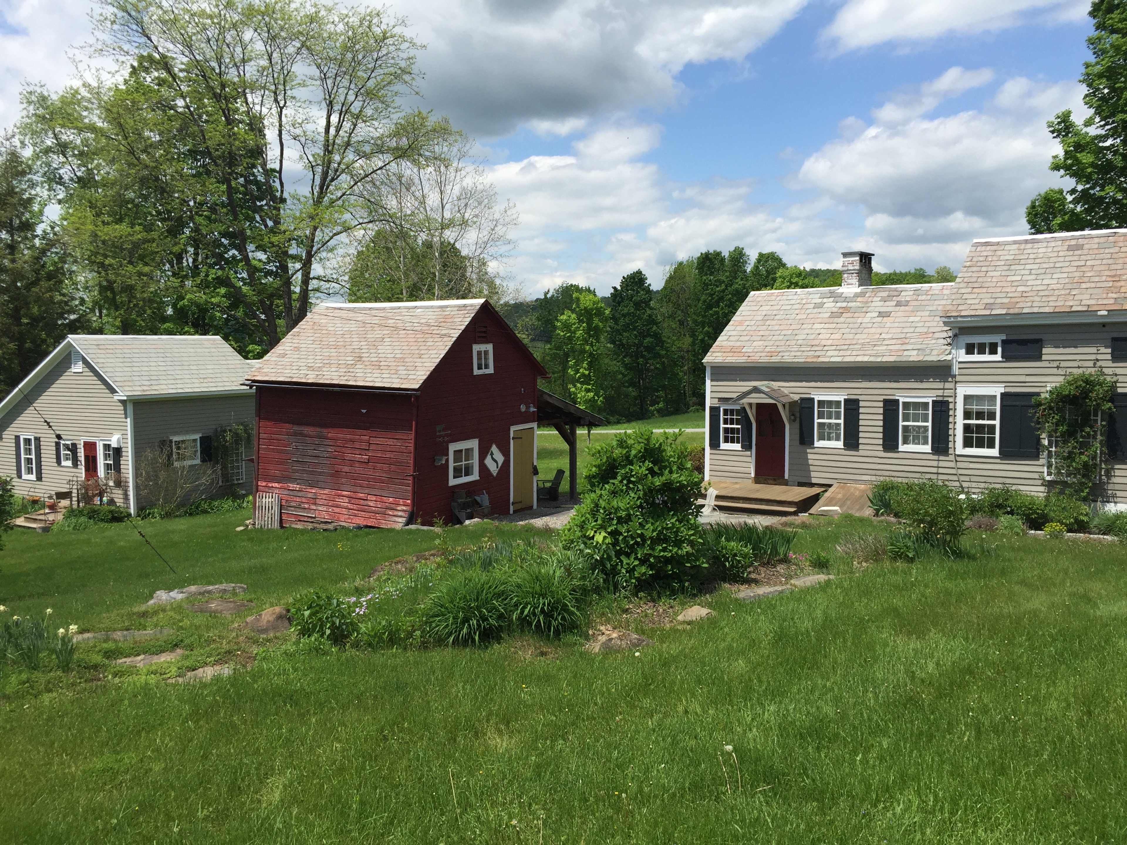 The image shows two distinct houses, one red and one gray, situated on a grassy landscape with trees in the background under a partly cloudy sky.