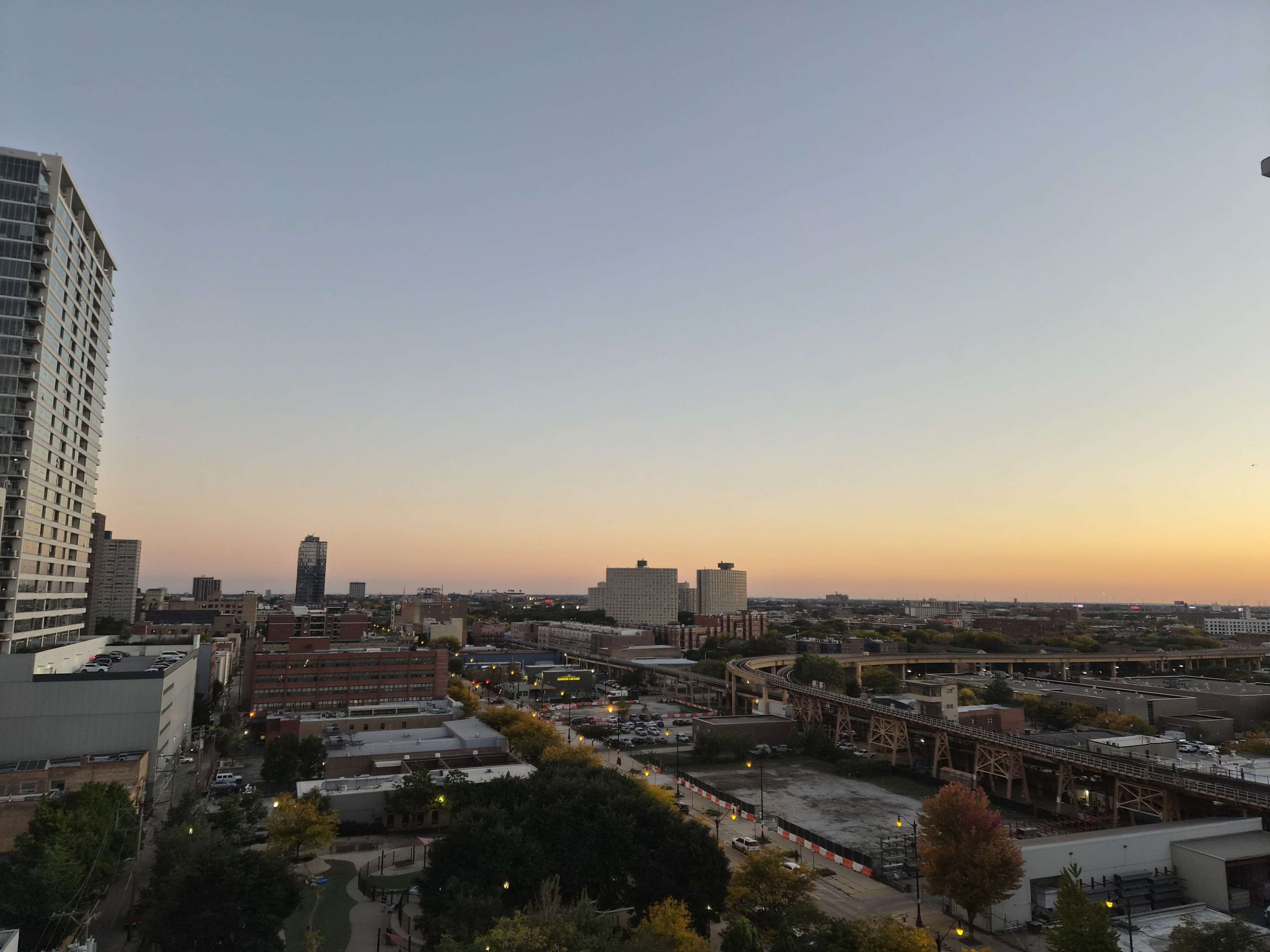 The image shows a skyline view of a city at sunset, with buildings, a highway, and a clear sky transitioning from daylight to twilight.