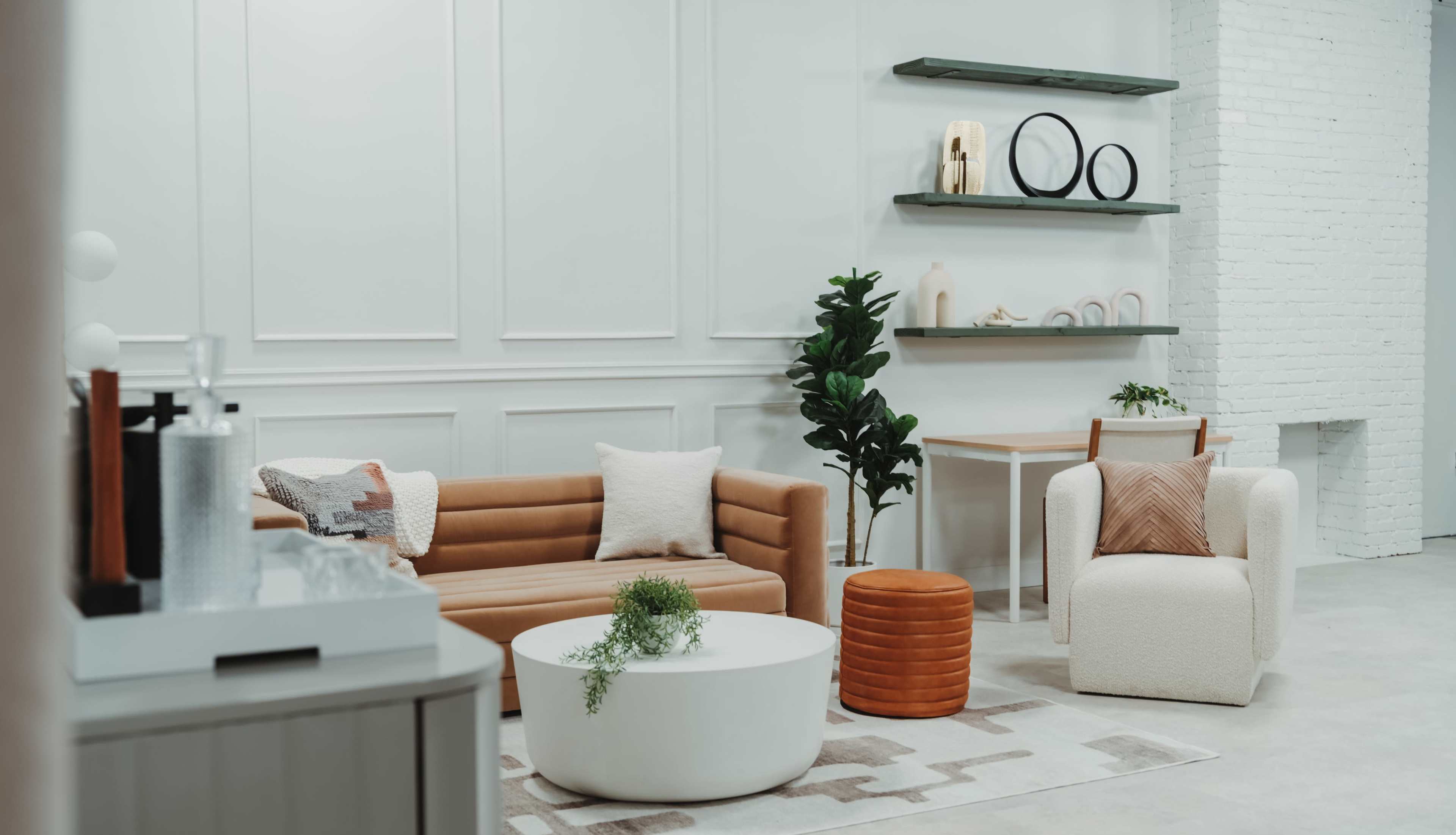 A modern living room features a brown sofa, a white circular coffee table, and a light-colored armchair, surrounded by minimalist decor and greenery.