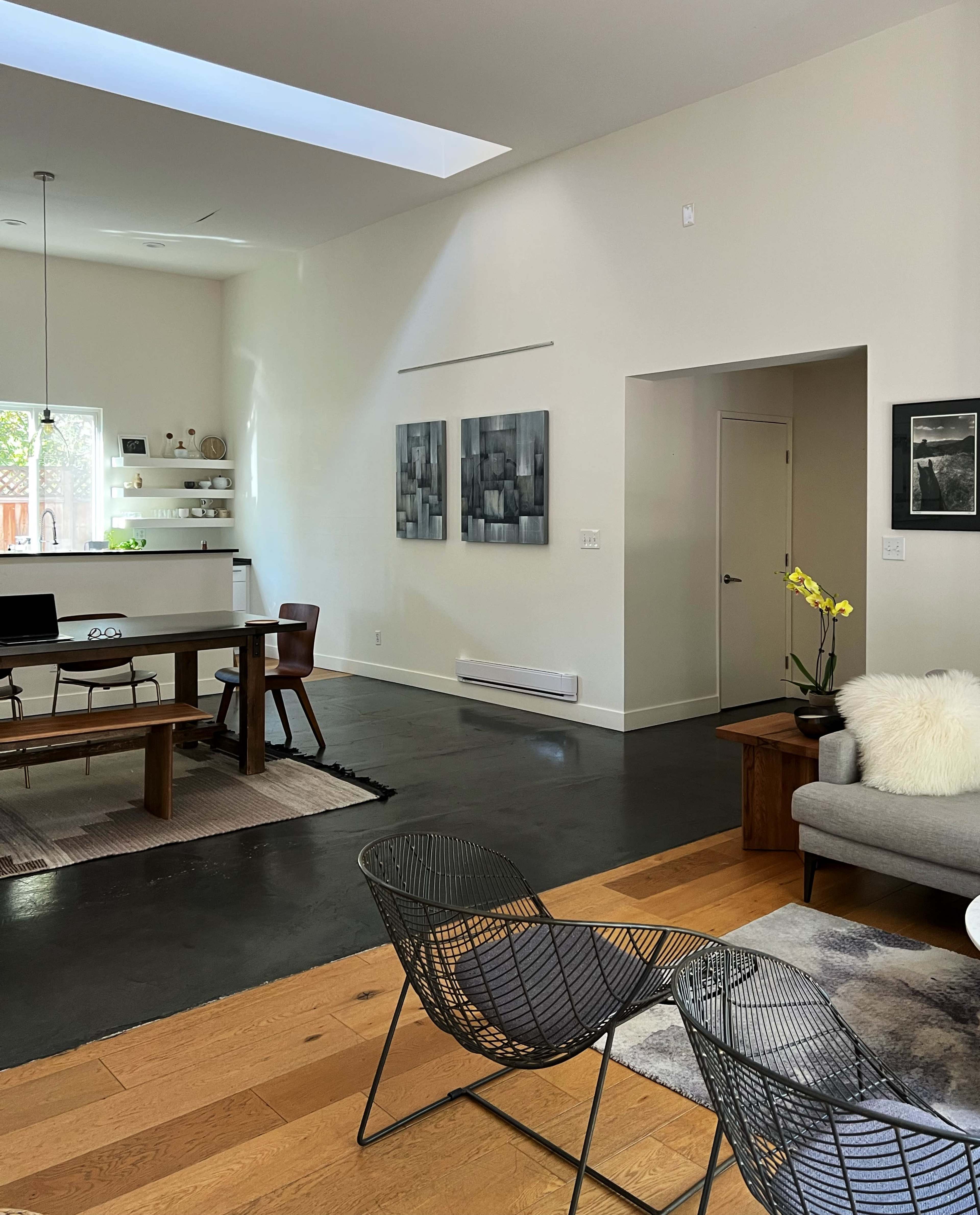 A spacious, modern living area with a dining table, two wire chairs, and minimalist decor, featuring large windows and artwork on the walls.