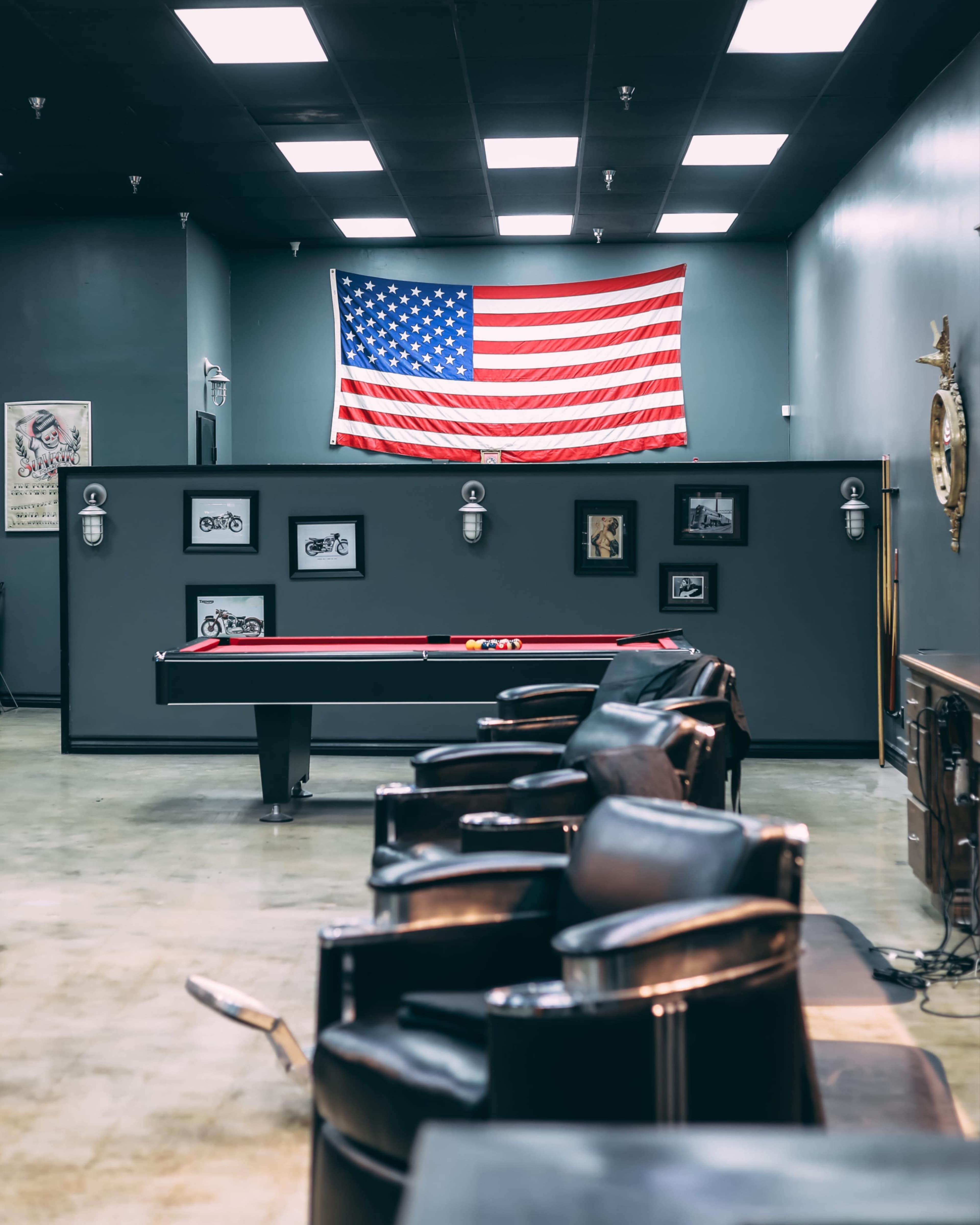 A barbershop interior featuring a pool table, leather chairs, and an American flag hanging on the wall.