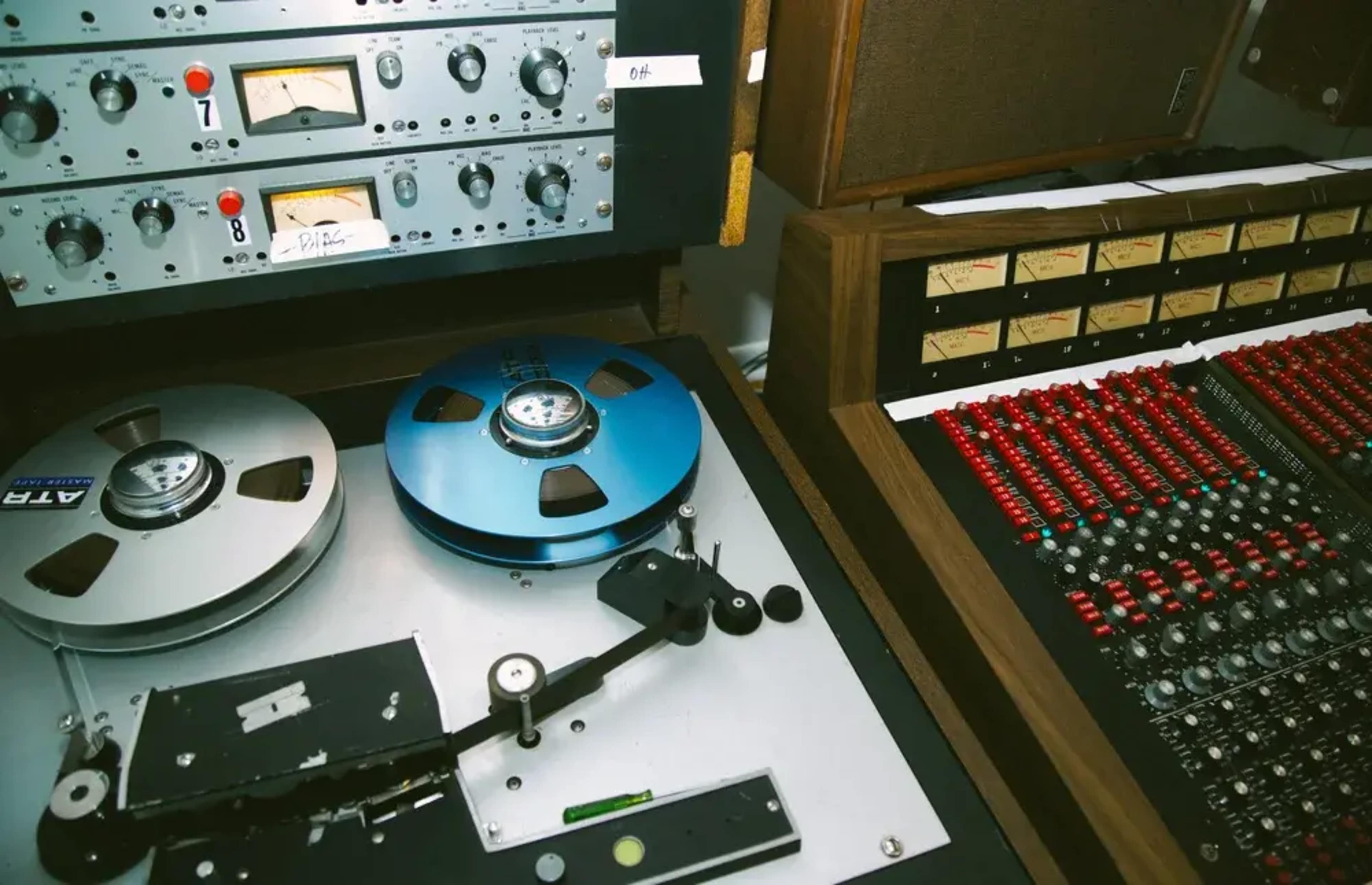 The image shows a vintage audio recording setup featuring reel-to-reel tape machines alongside a control console with numerous red knobs and faders.