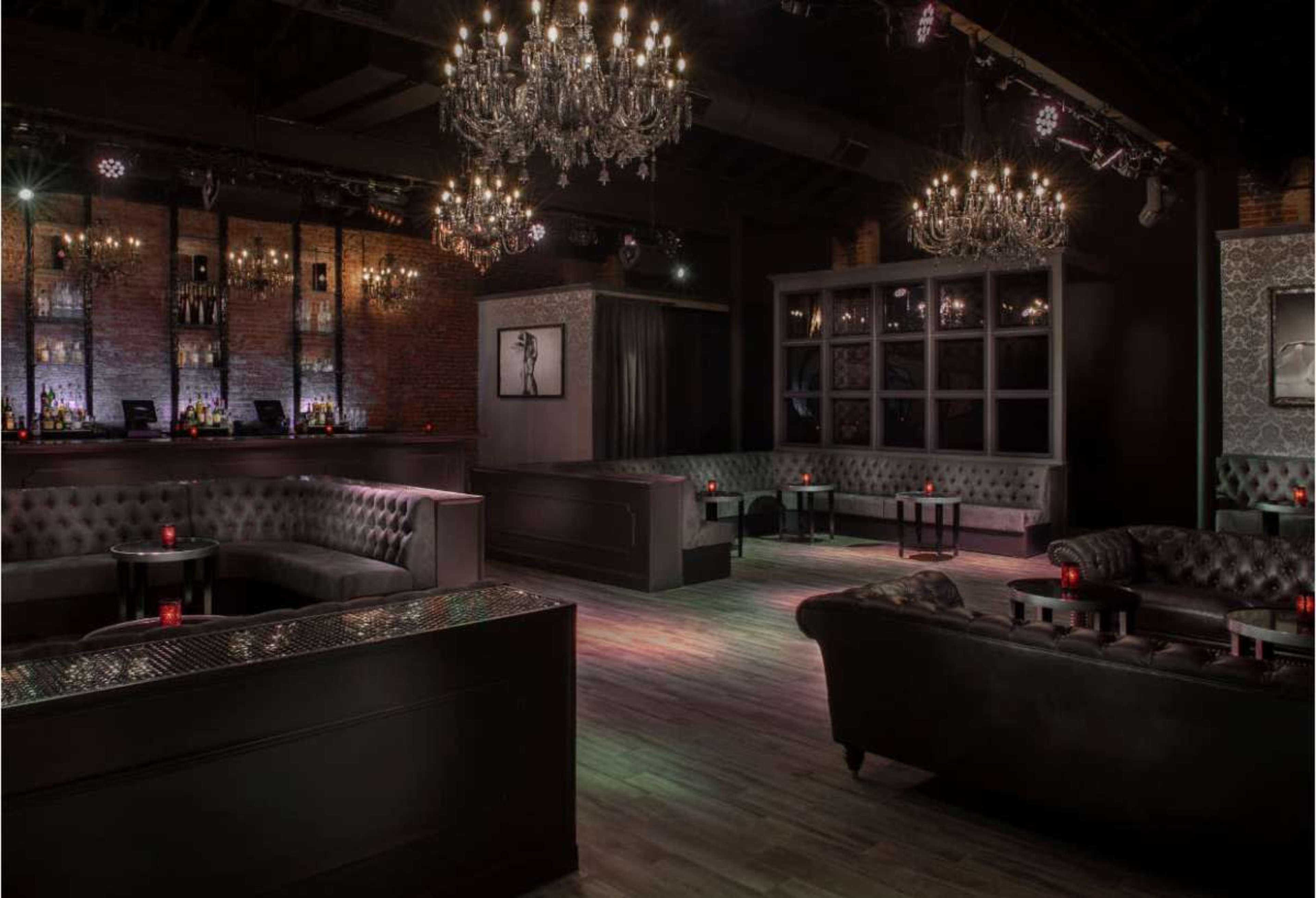 The image shows a dimly lit lounge featuring plush seating, chandeliers, and a bar area with bottles displayed against a brick wall.