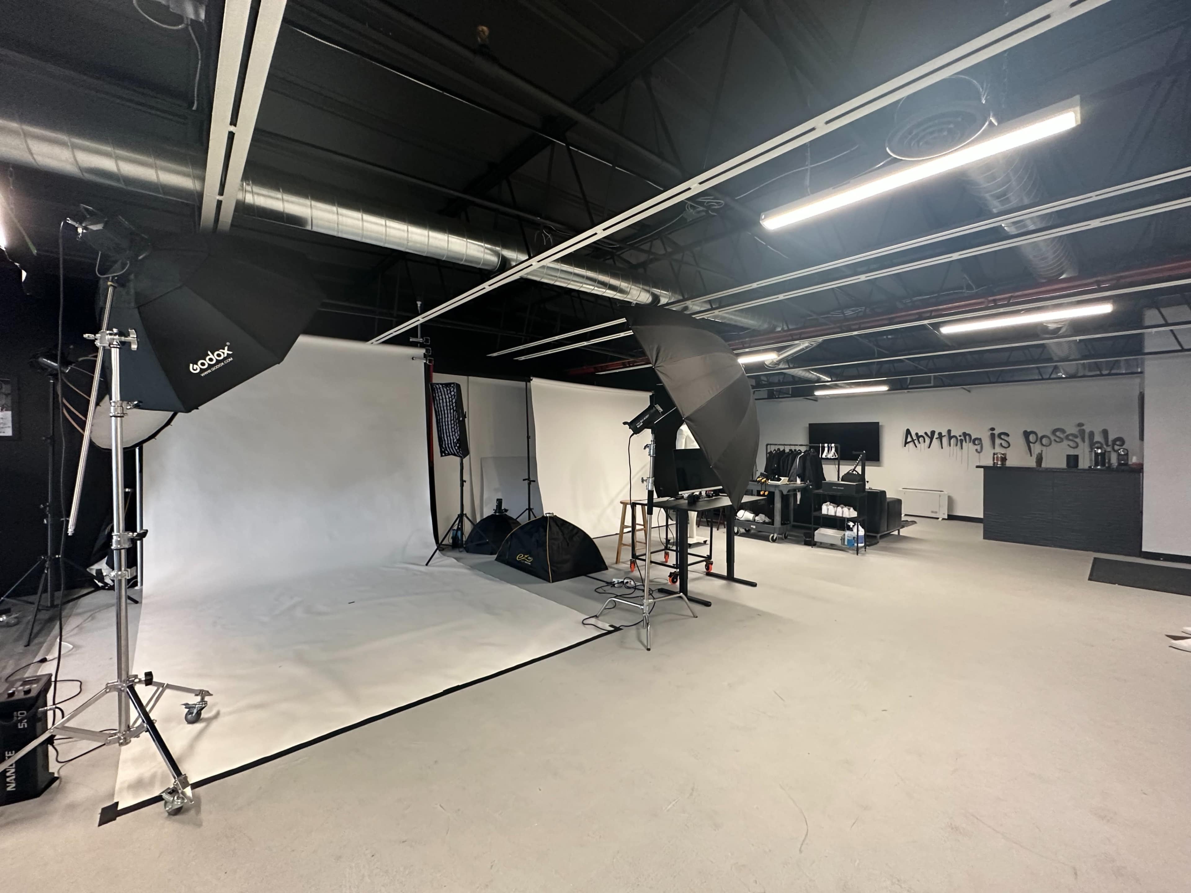 A photography studio equipped with lighting equipment, a blank backdrop, and a minimalistic workspace with a wall displaying the phrase "Anything is possible."