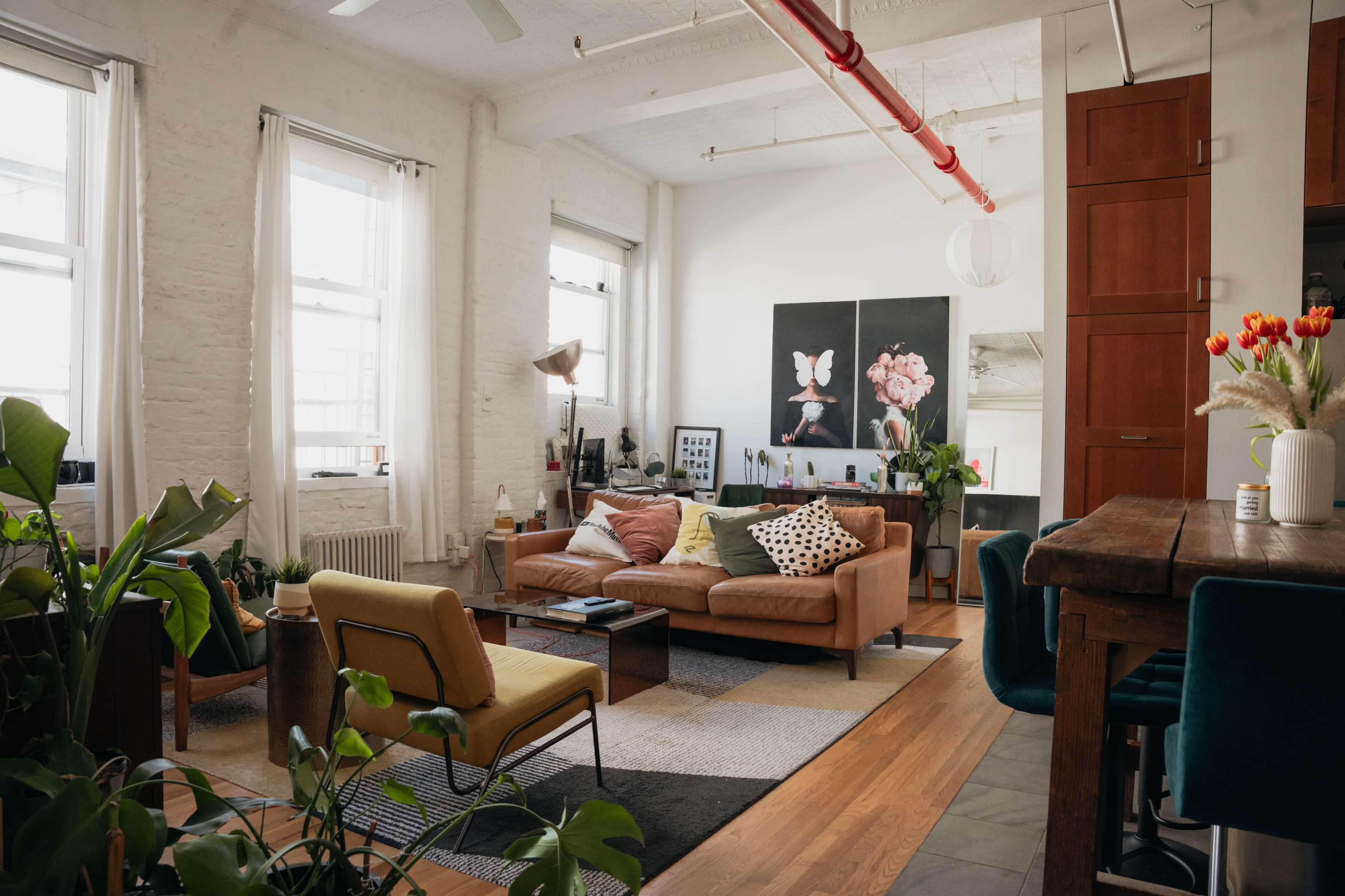A modern living room features a brown leather sofa, colorful cushions, a wooden dining table, and plants throughout, with large windows allowing natural light to fill the space.