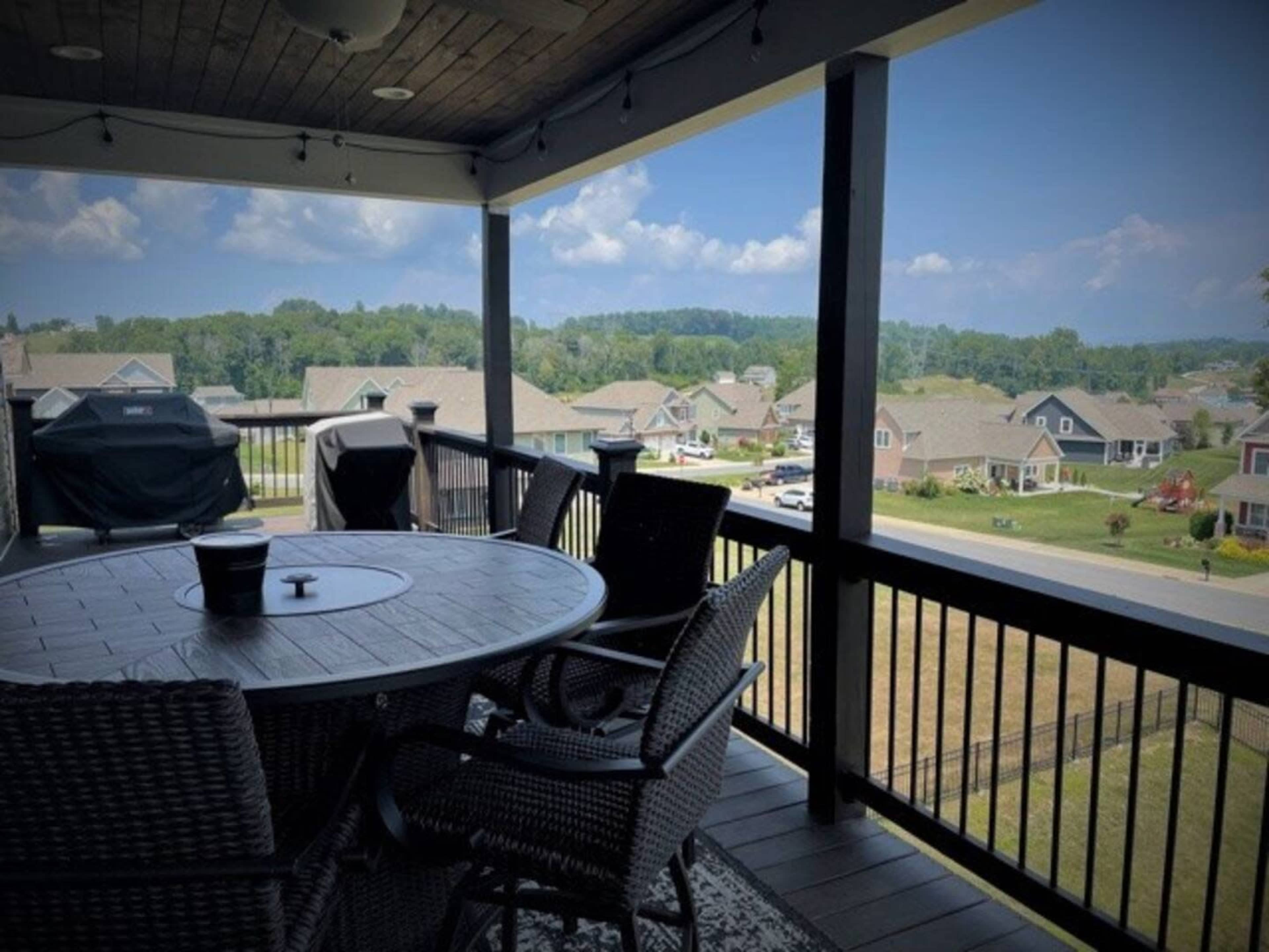A covered balcony features a round table and chairs with a view of suburban homes and greenery in the background.