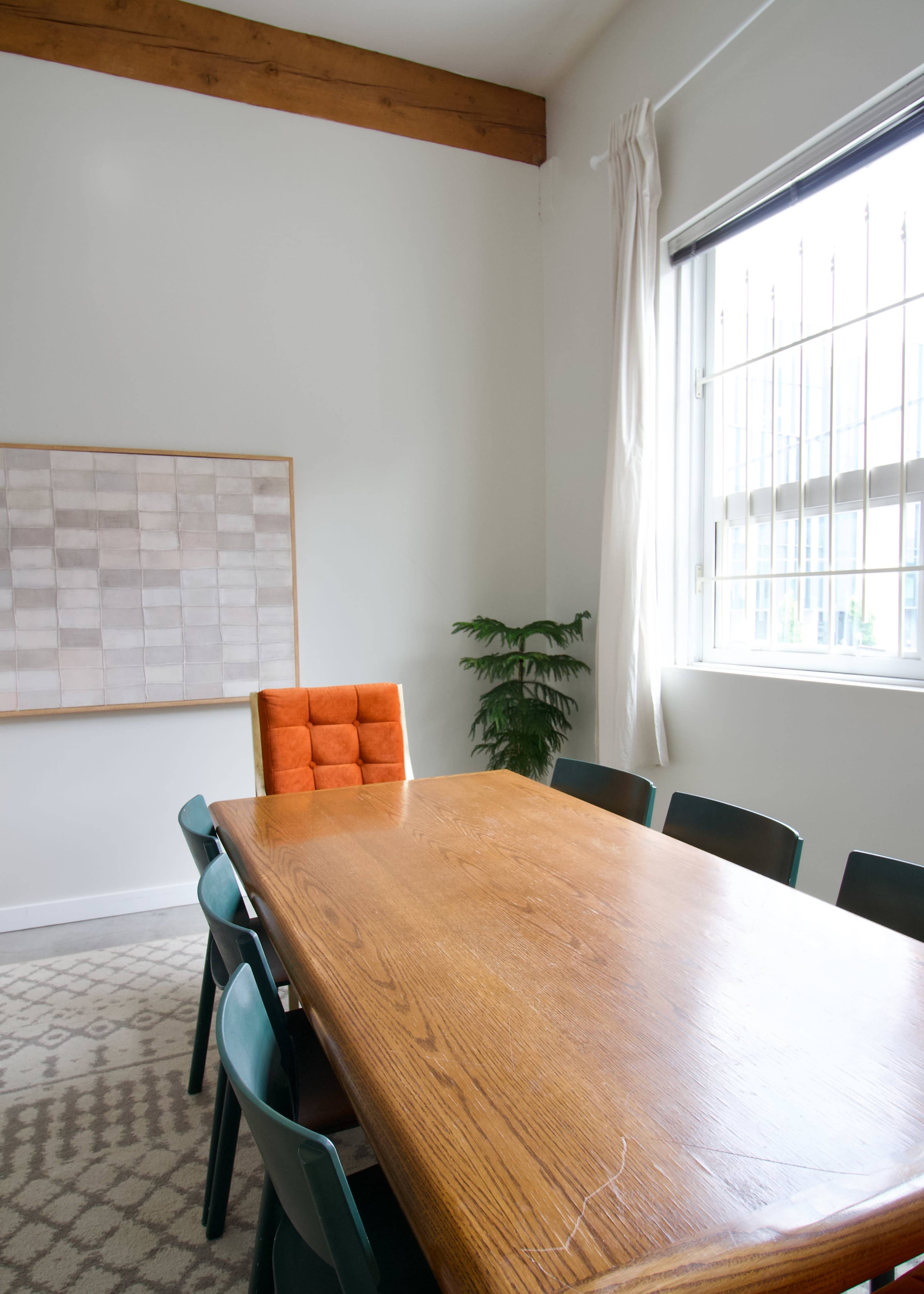 A large wooden table with a row of green chairs is positioned in a bright room featuring a window with bars, an orange chair, and a decorative plant.