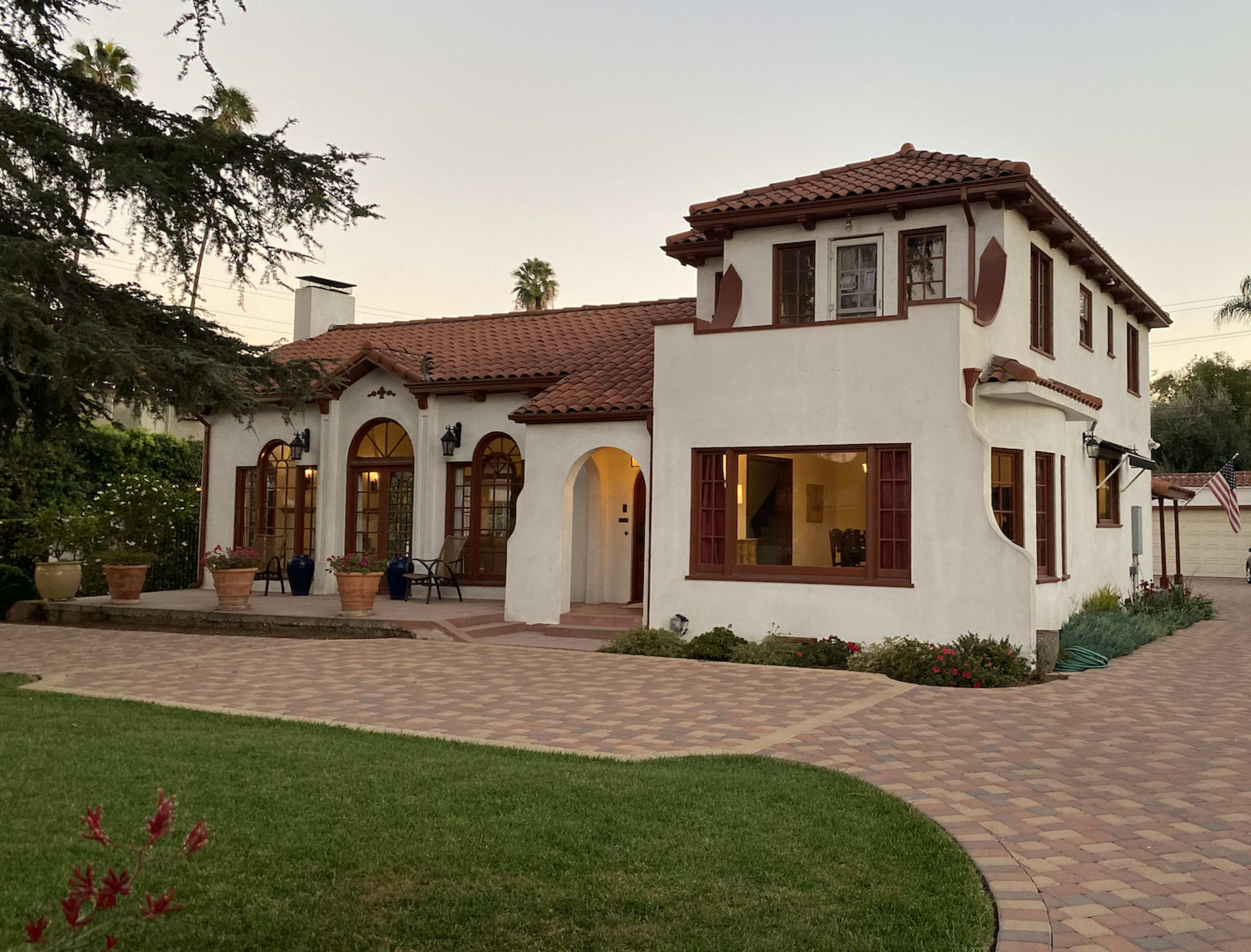 A stucco house with a red tile roof features arched windows and a brick path leading to a landscaped yard.