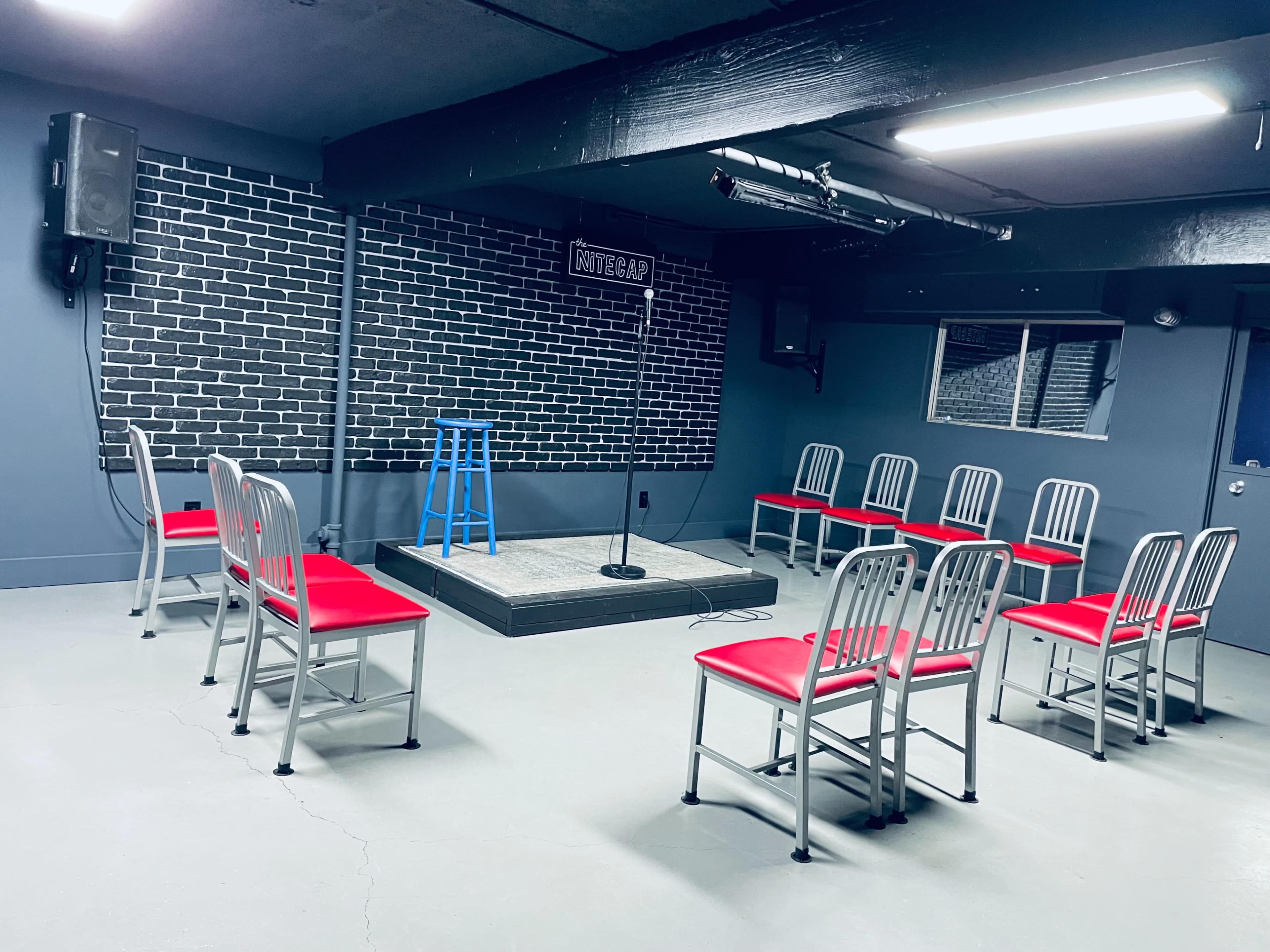 A small performance space features a circular arrangement of red and silver chairs facing a raised platform with a microphone, against a backdrop of black brick walls.